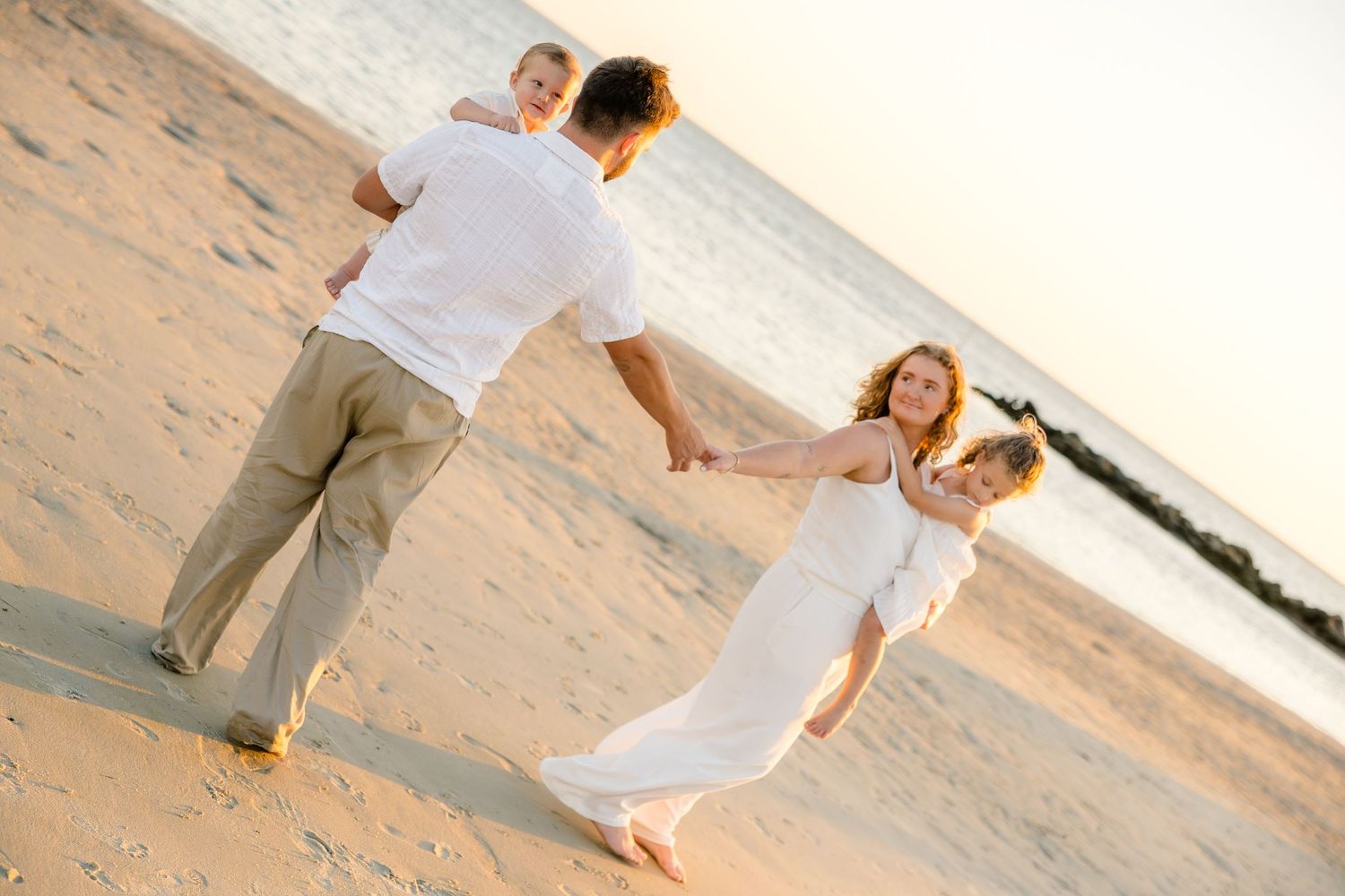 Happy family of four enjoying a sunny beach walk, parents in white outfits swinging children on golden sandy shore.