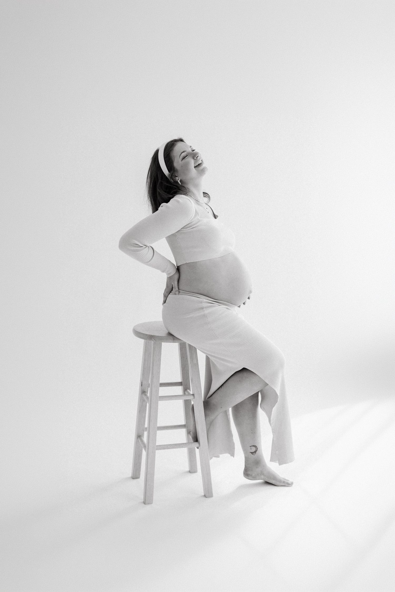 Black and white maternity photo of a smiling pregnant woman sitting on a stool in a white dress.