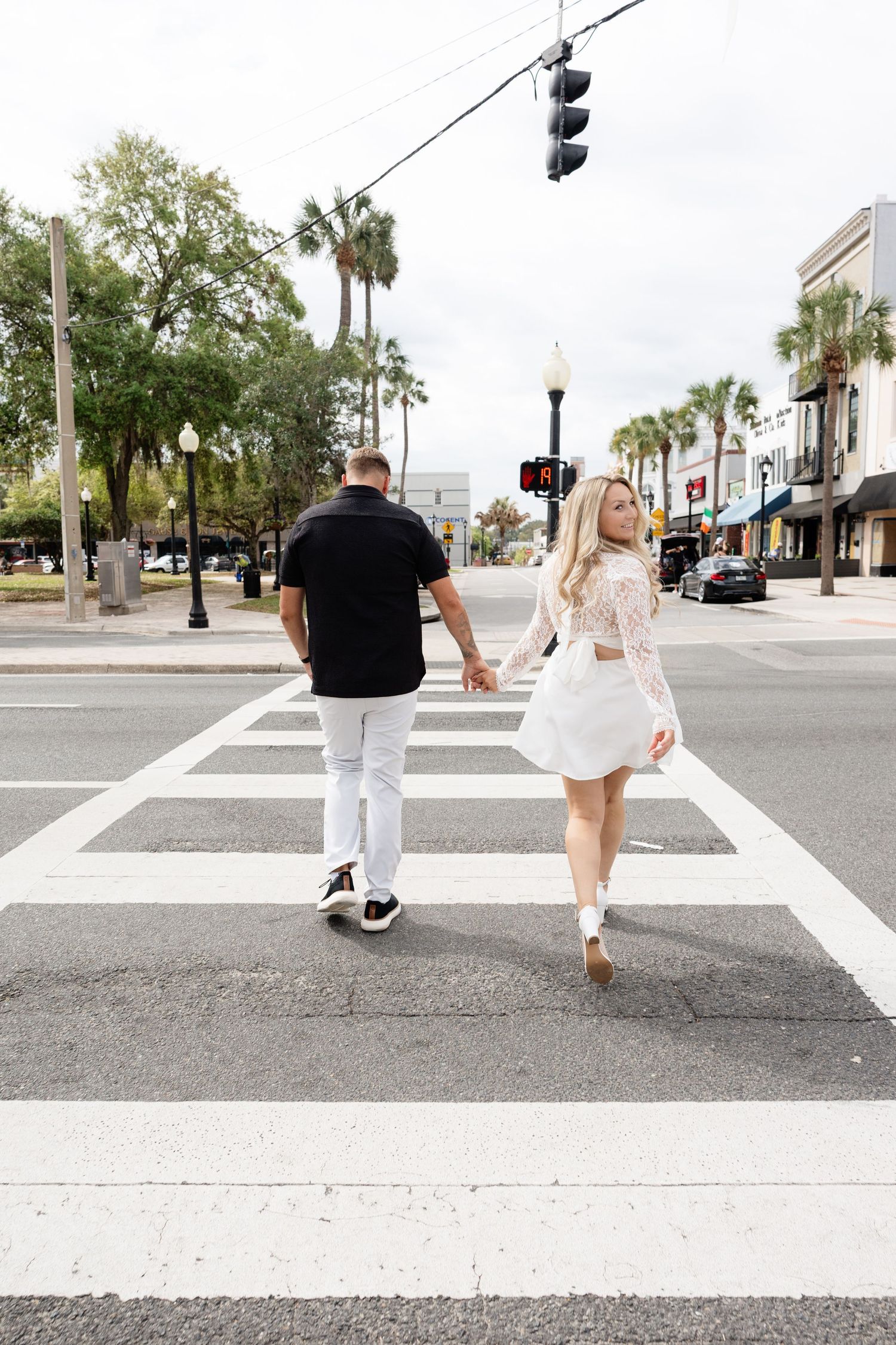 Couple holding hands while crossing a zebra crossing on a sunny urban street, woman in white dress looks back.
