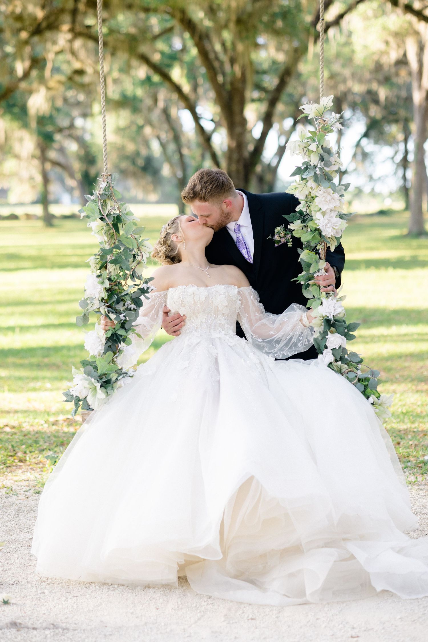 Bride and groom sharing a kiss on a floral swing adorned with greenery and white flowers in a lush outdoor park setting.