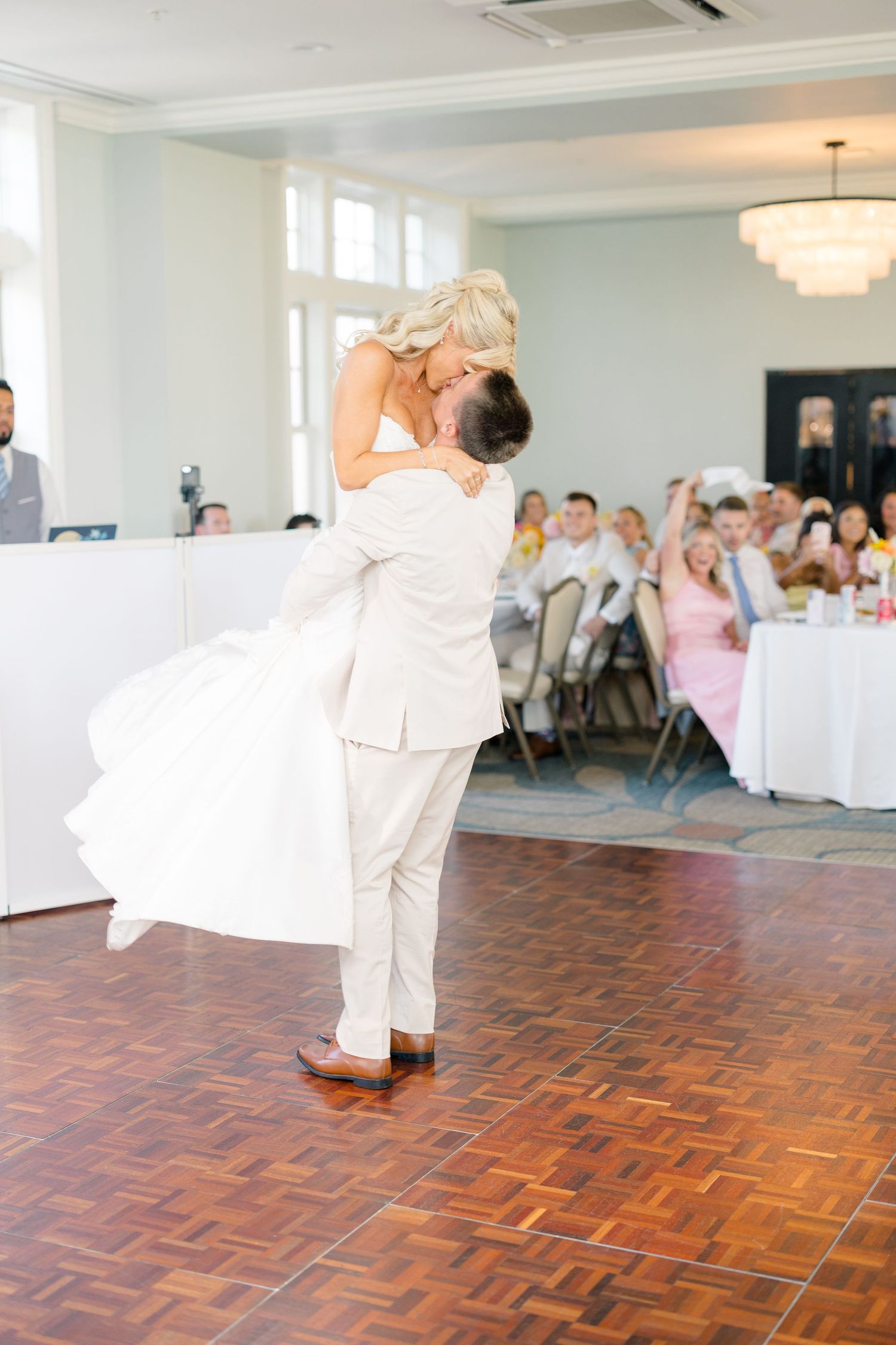 Groom lifts bride during romantic first dance at elegant wedding reception with guests watching in background.
