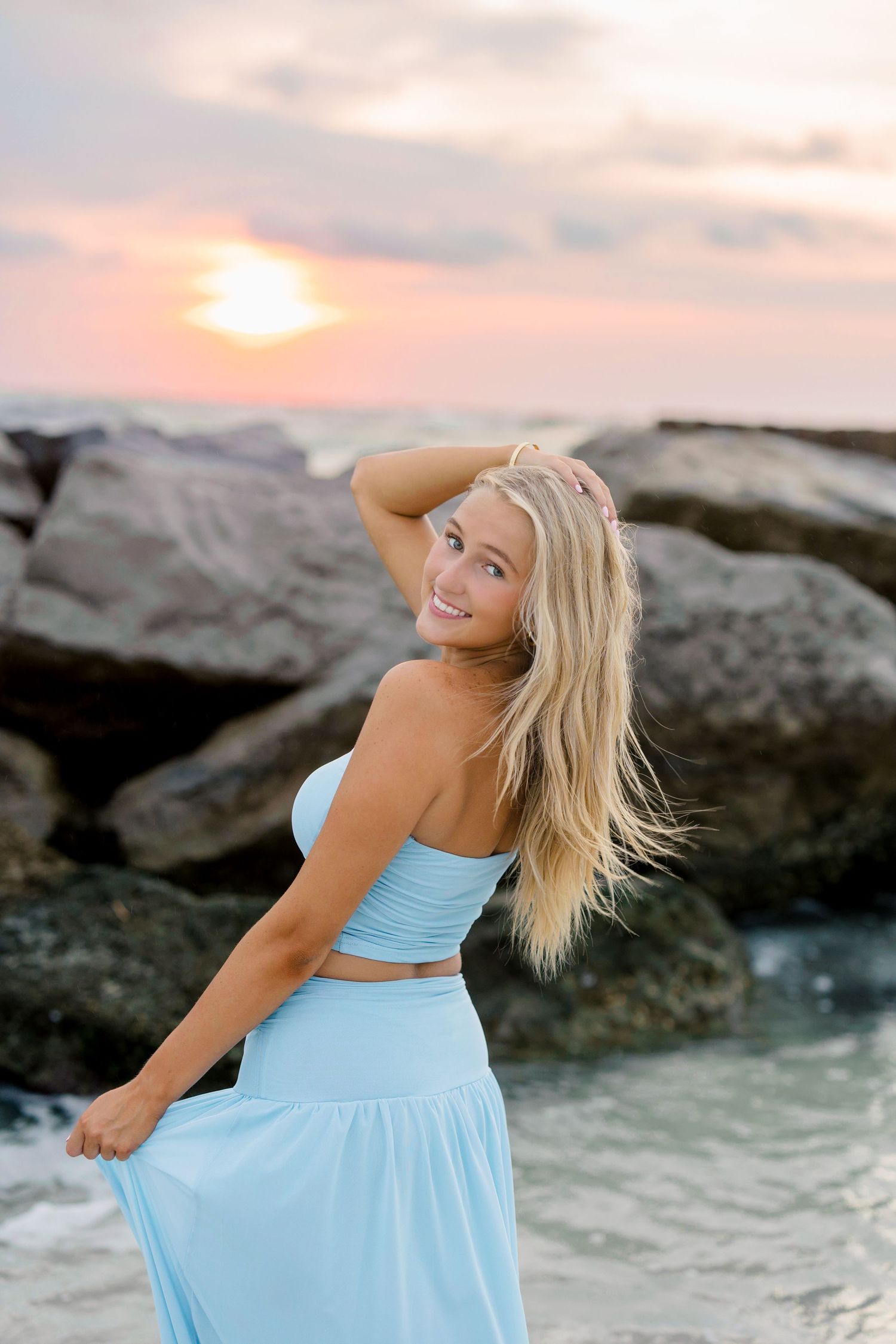 Blonde woman in light blue crop top and maxi skirt poses on rocky beach at sunset, smiling over shoulder.