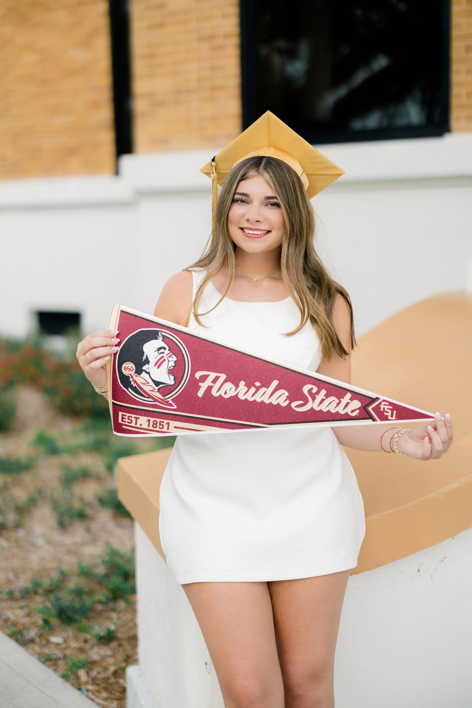 A smiling female graduate in a white dress and gold cap holds a Florida State University pennant outdoors.