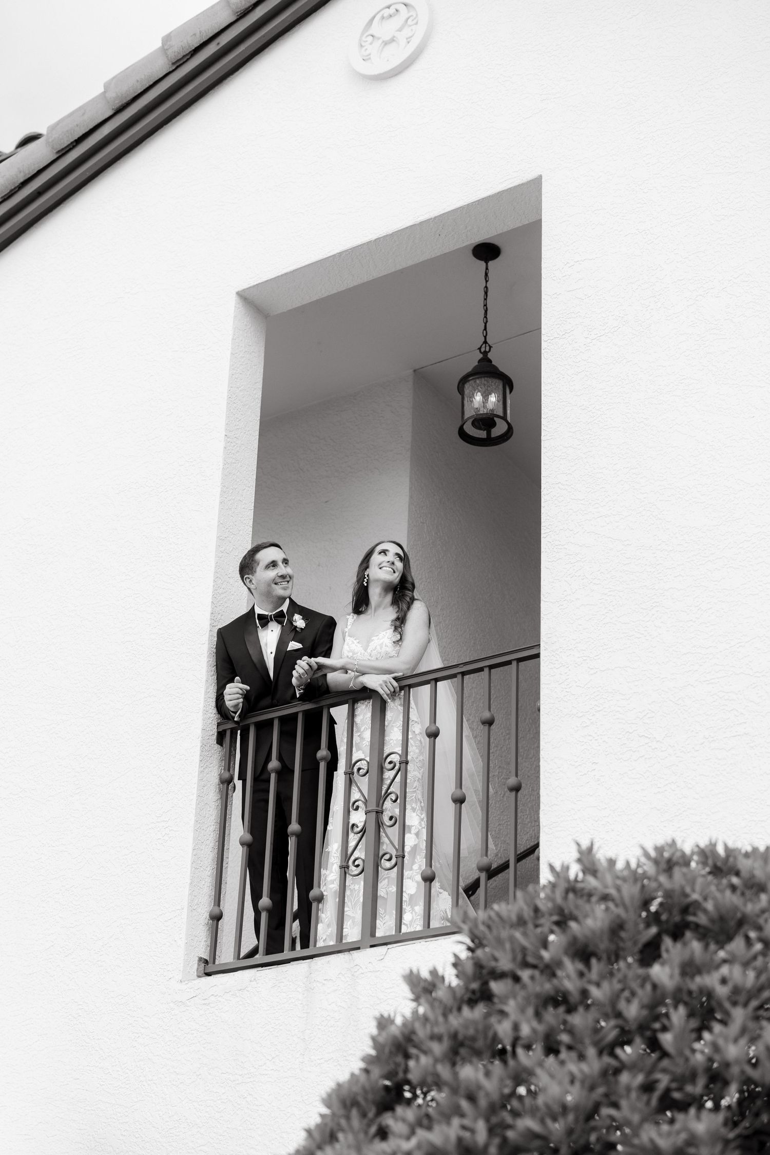 Black and white photo of bride and groom standing on ornate balcony of white stucco building.