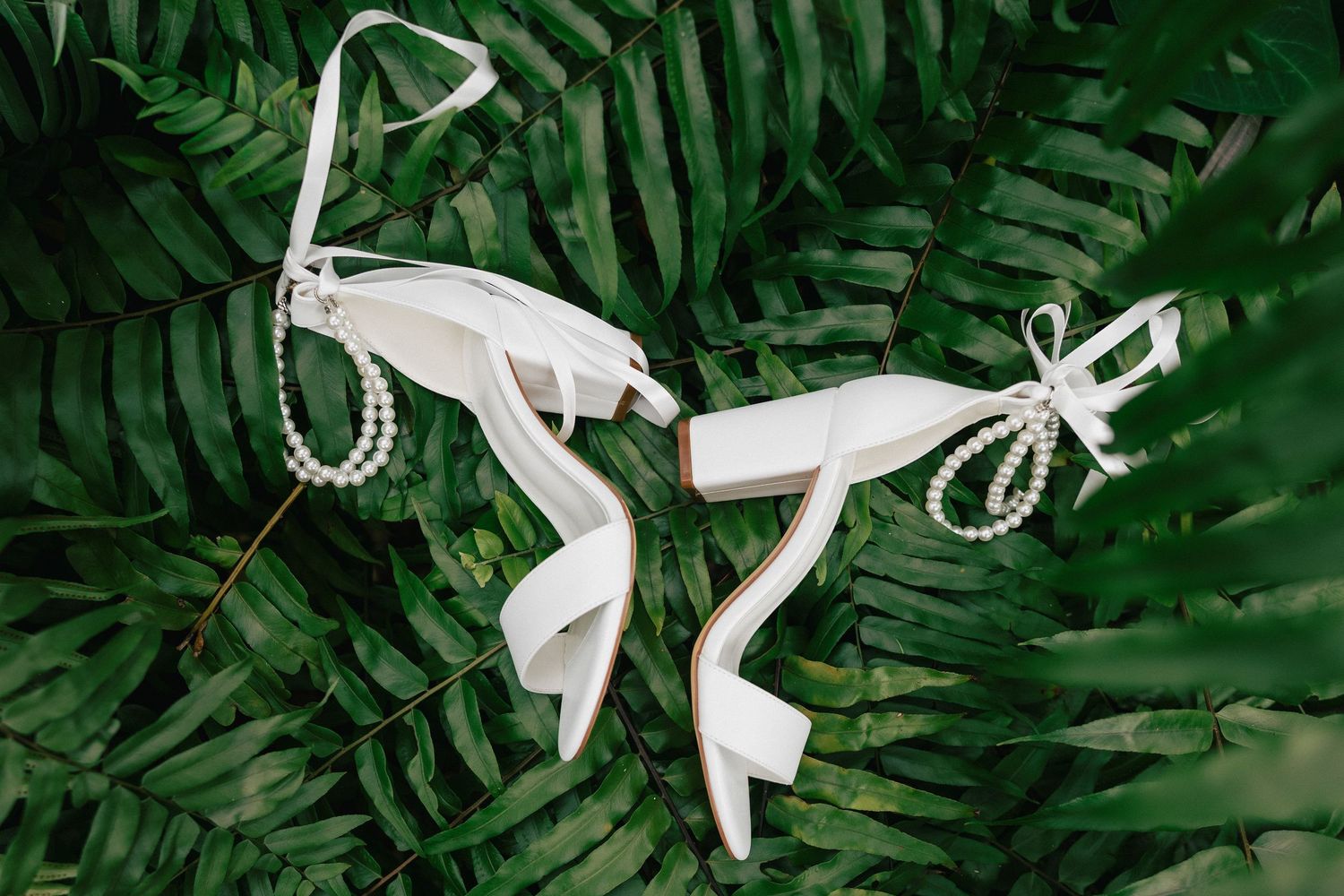 White bridal heels with pearl ankle straps displayed on lush green fern leaves for a wedding.