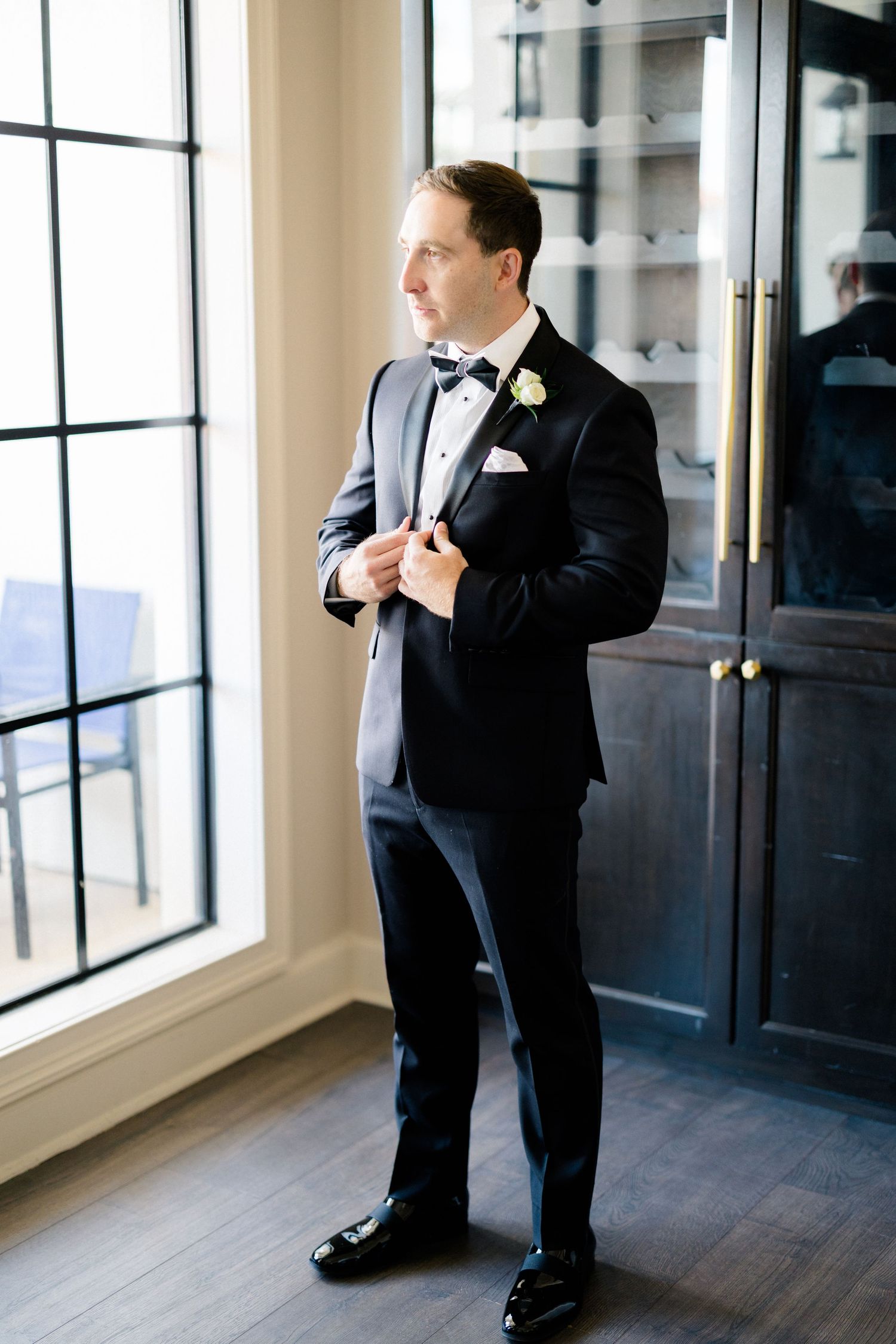 Groom in black tuxedo with bow tie and white boutonniere adjusting jacket by window.