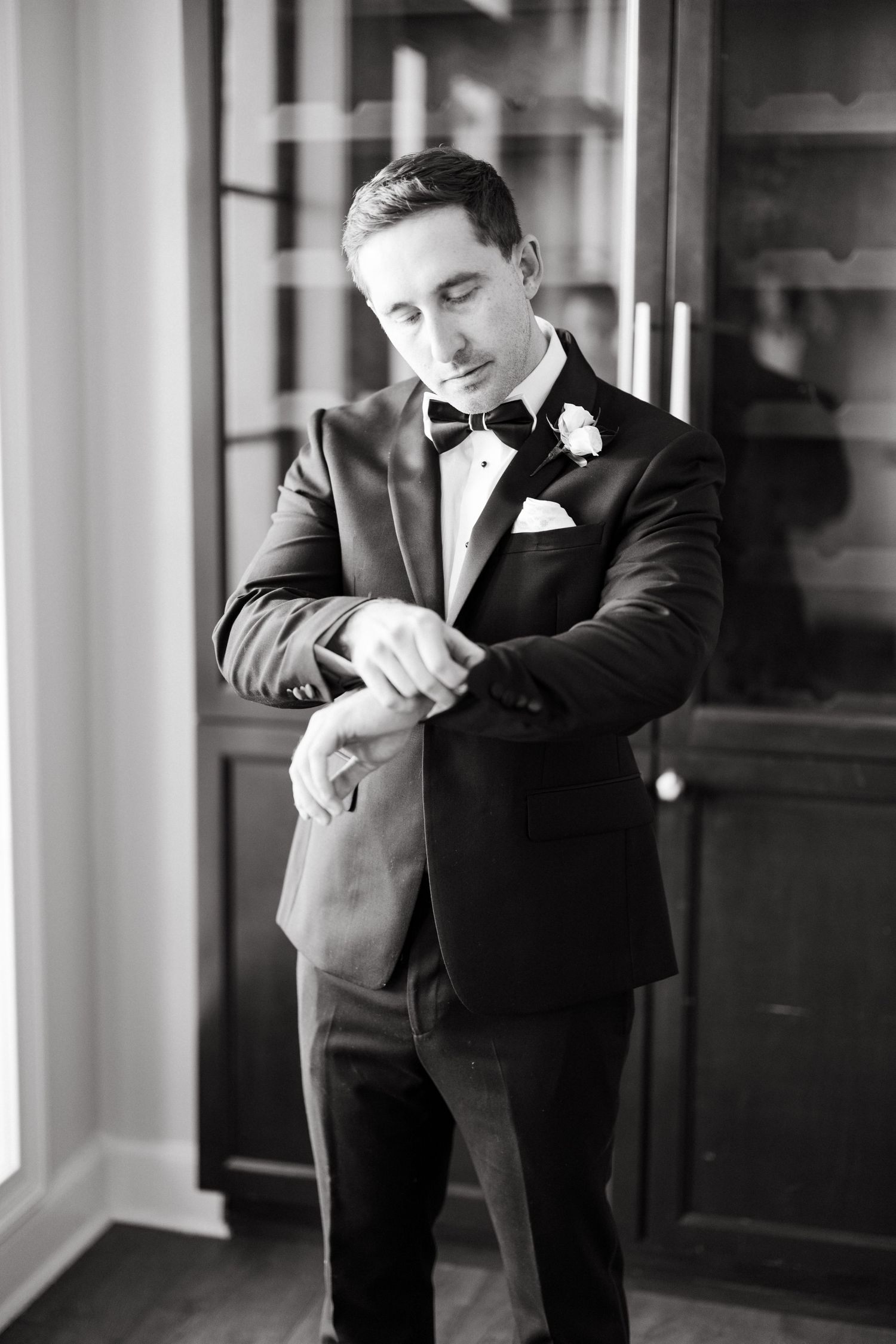 Groom in black tuxedo with bow tie and boutonniere adjusting his cufflinks in elegant black and white photo.