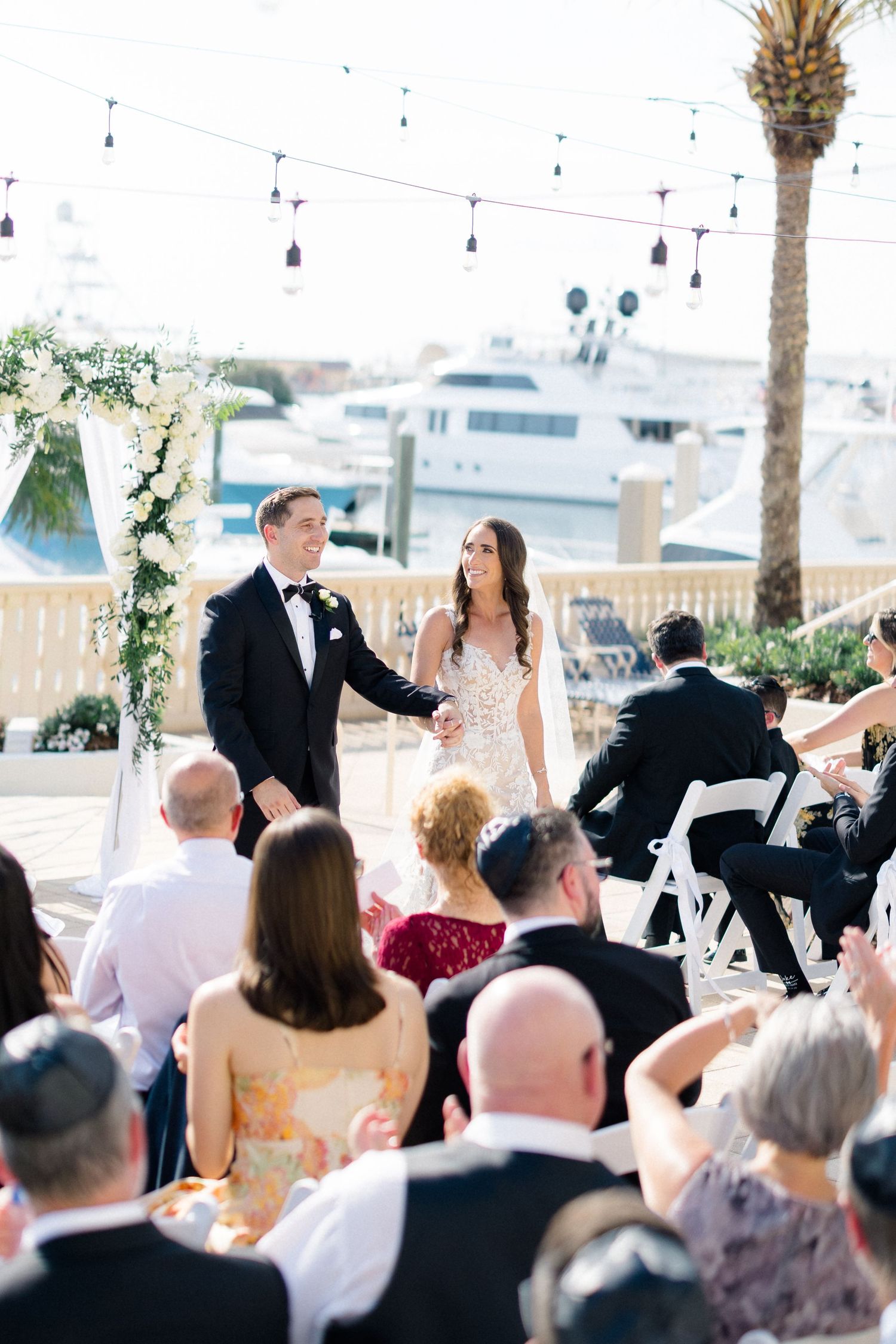 Bride and groom hold hands at outdoor waterfront wedding ceremony with floral arch and string lights.