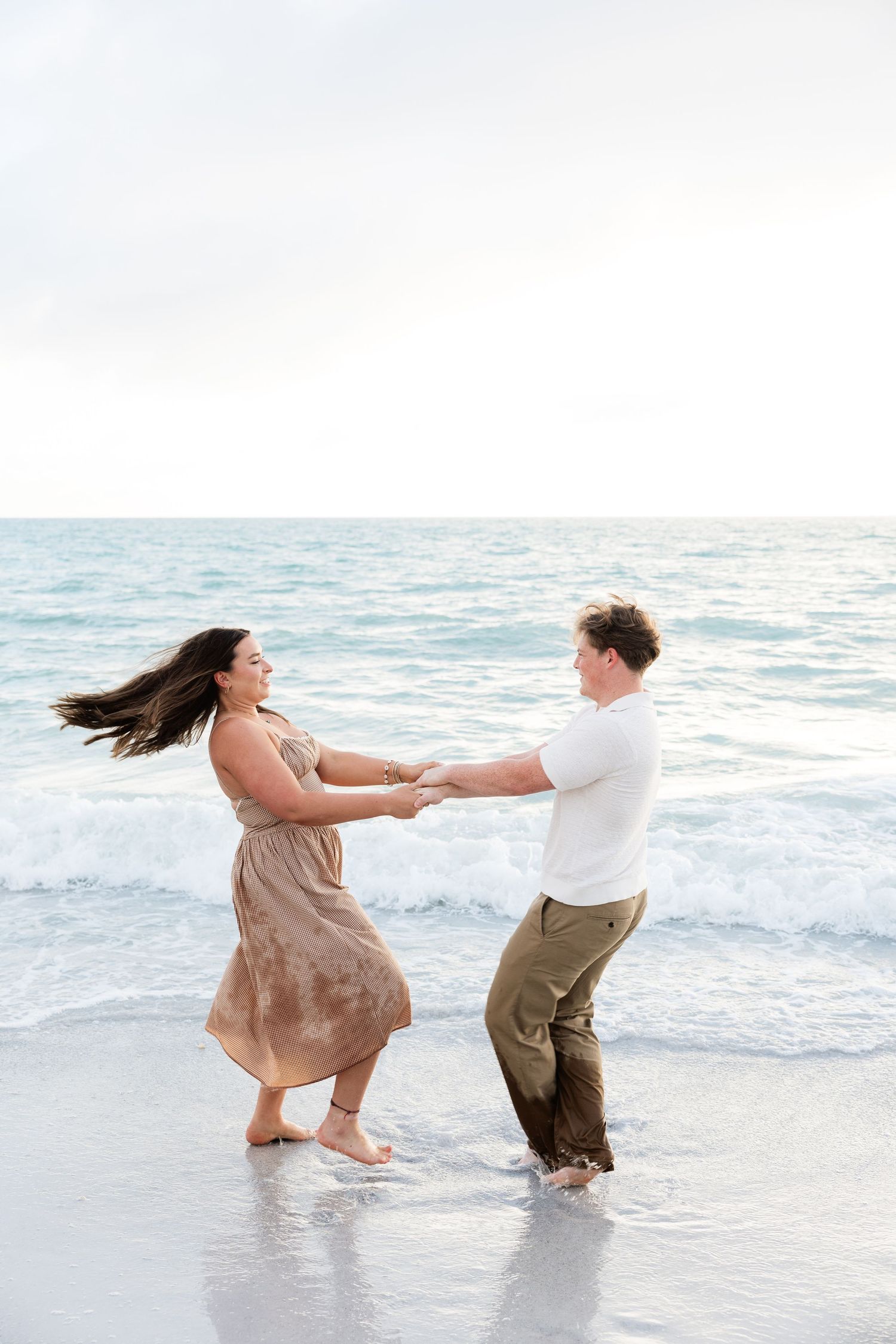 A couple dances joyfully on a beach shoreline, holding hands with ocean waves behind them.