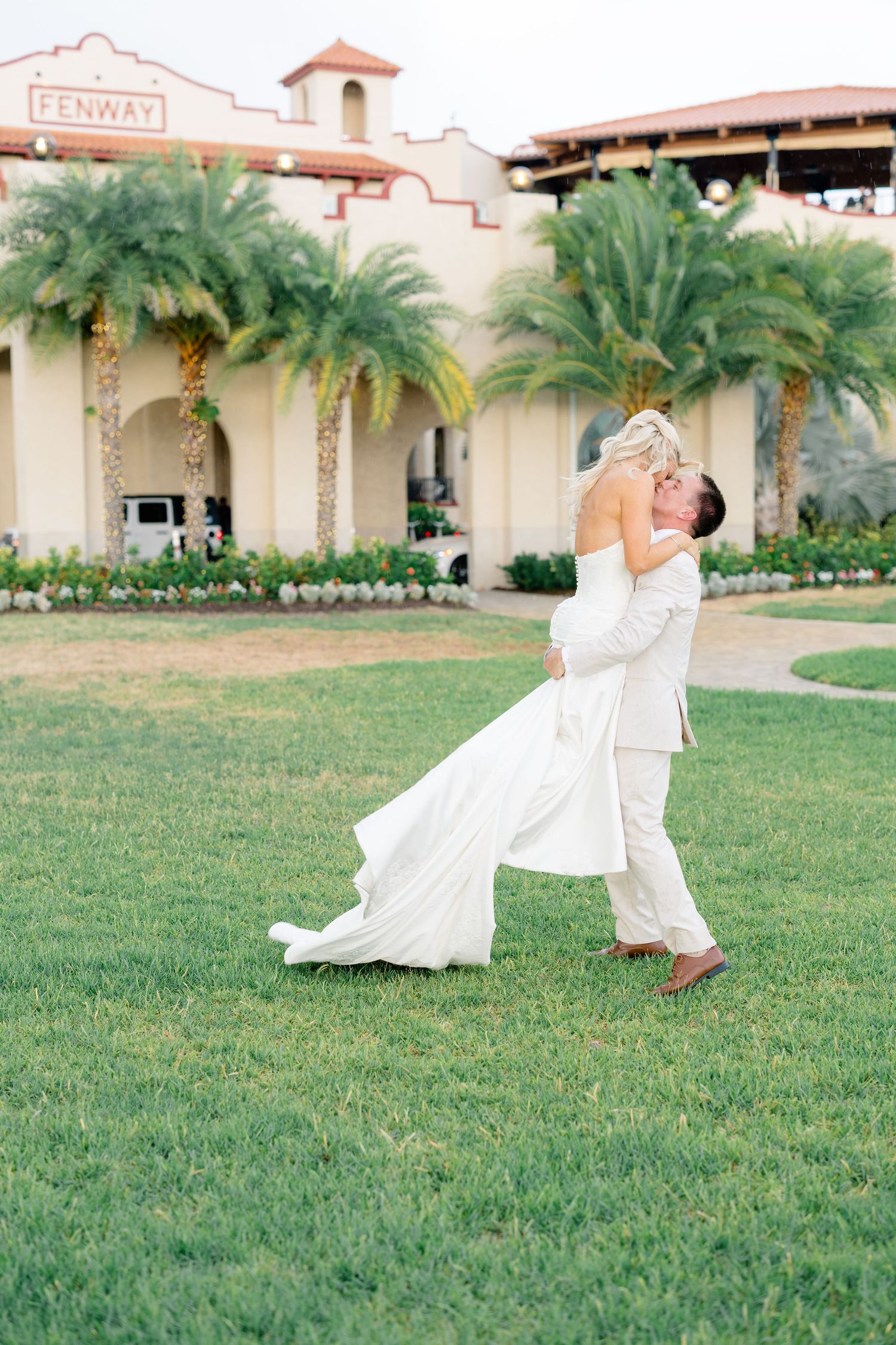 Groom lifting bride in white dress on green lawn outside Fenway venue with palm trees.