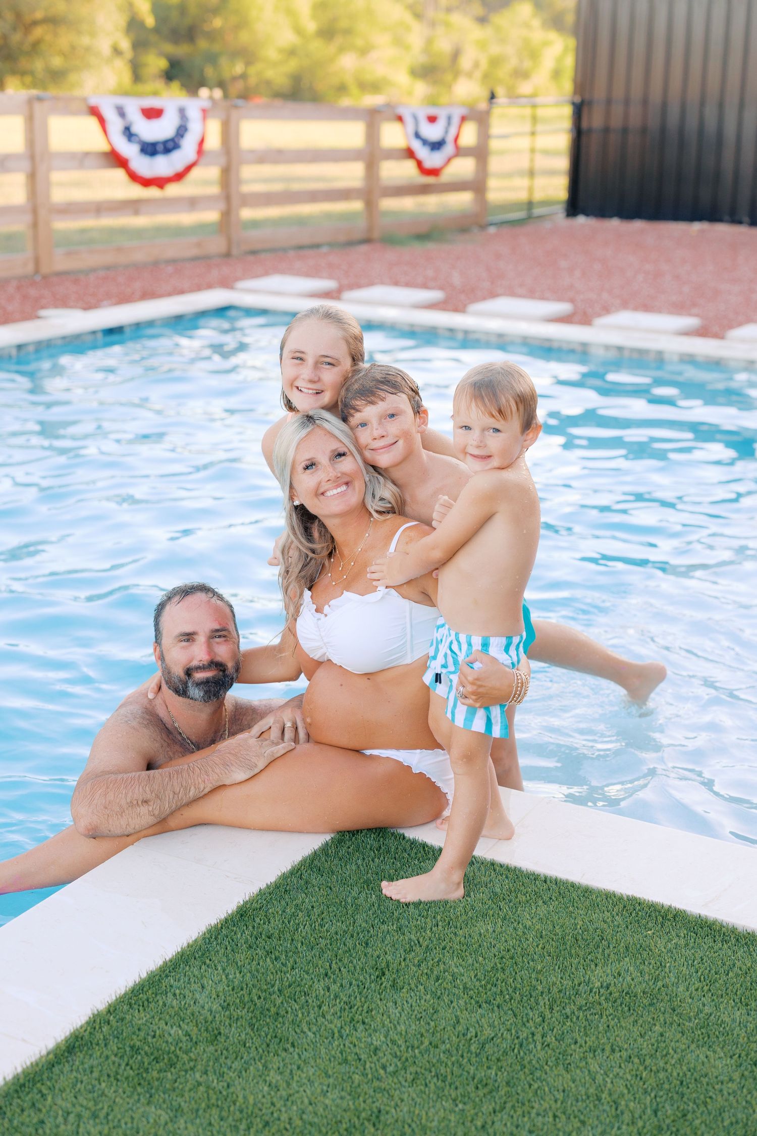 Happy family of five posing together at a backyard pool with patriotic bunting decorations on a sunny day.
