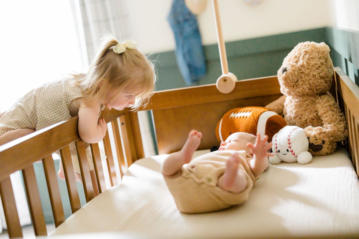 Toddler girl leaning over wooden crib looking at newborn baby sibling with teddy bear toys nearby.