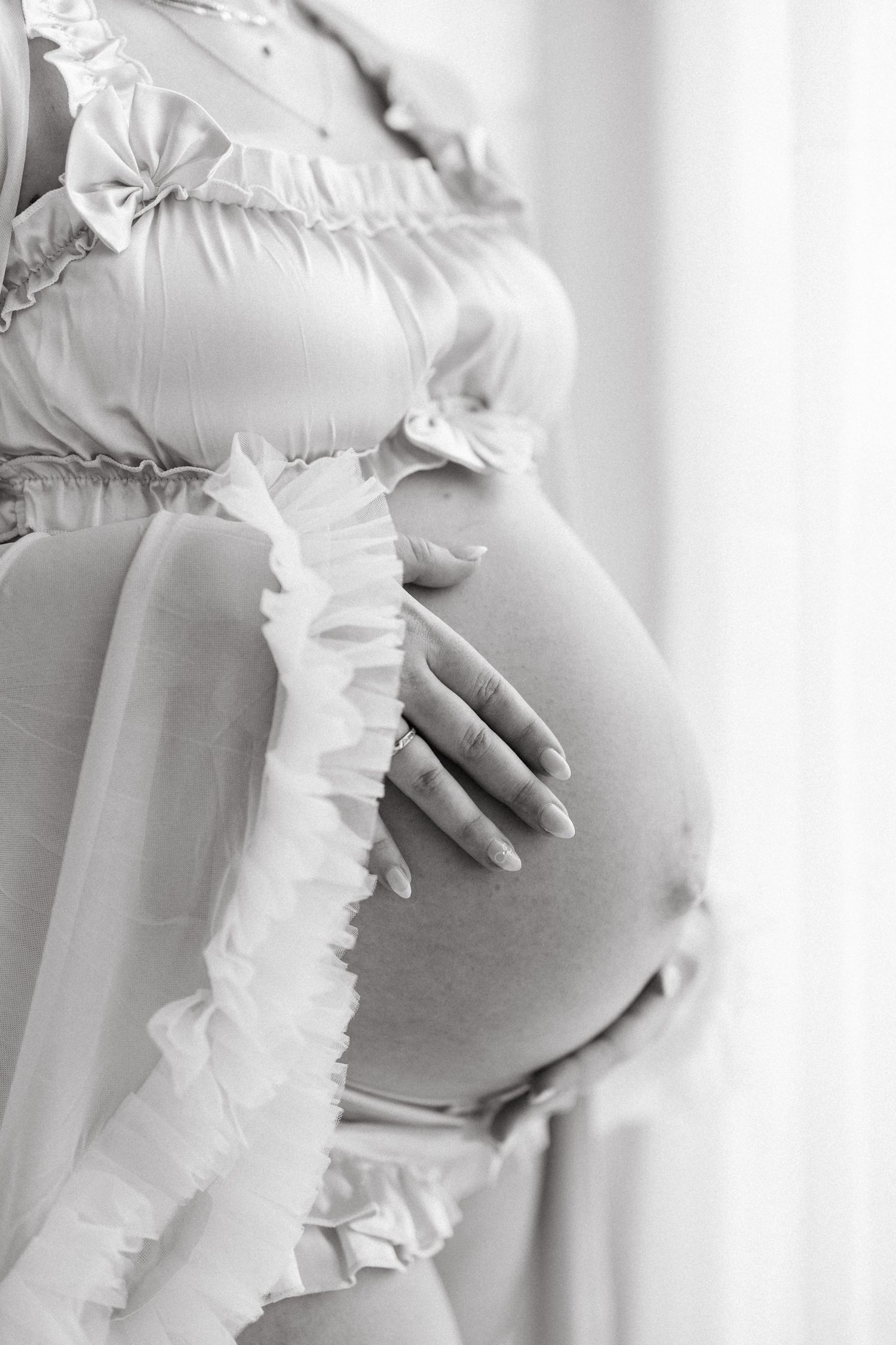 Black and white photo of a pregnant woman in a ruffled outfit cradling her bare baby bump.
