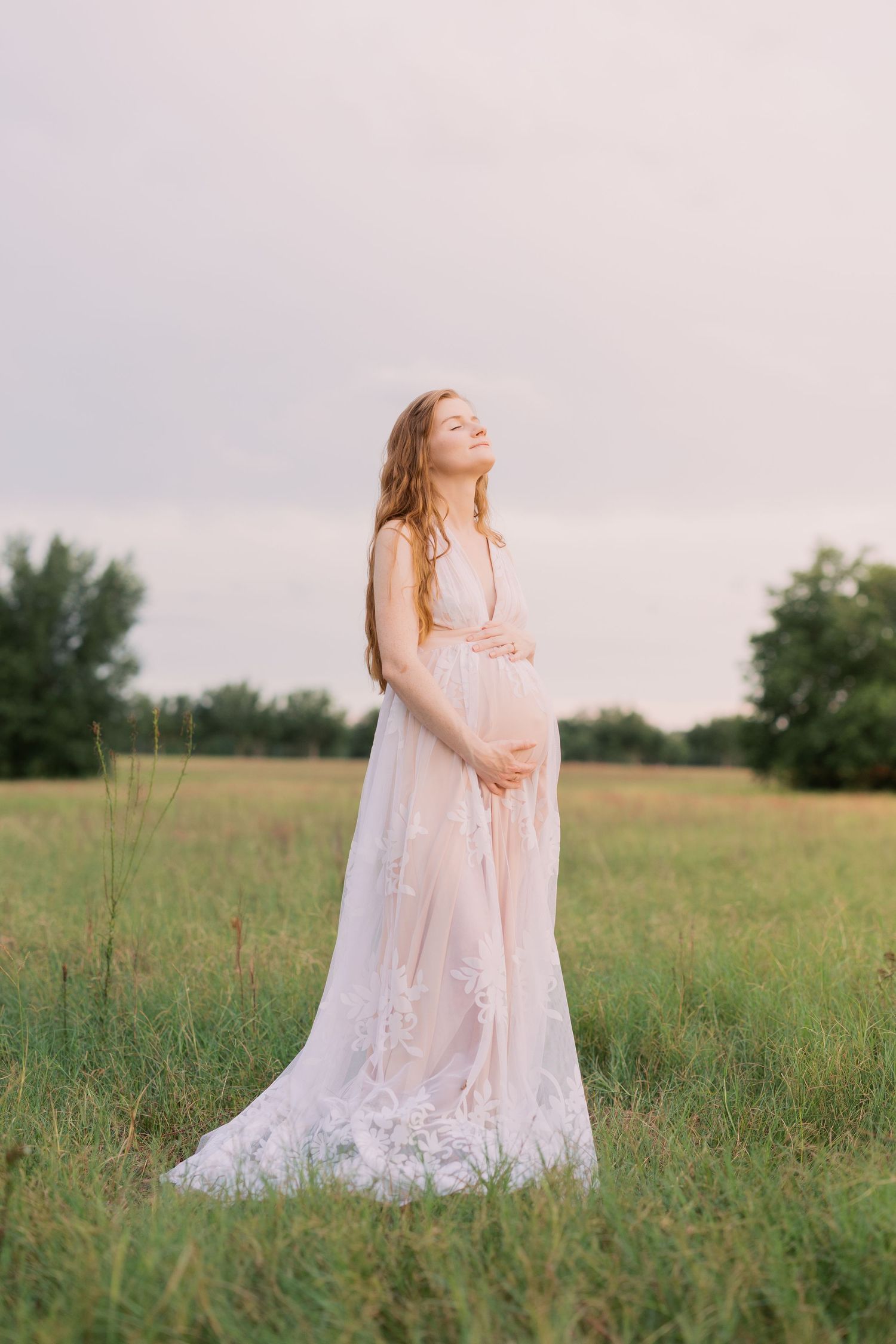 Pregnant woman in sheer white lace gown standing in green field, eyes closed, hands on belly at sunset.