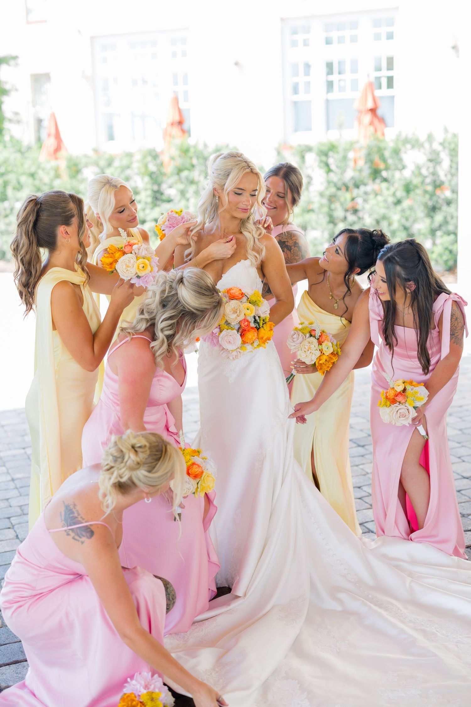 Bride surrounded by bridesmaids in pink and yellow dresses holding colorful bouquets at an outdoor wedding ceremony.