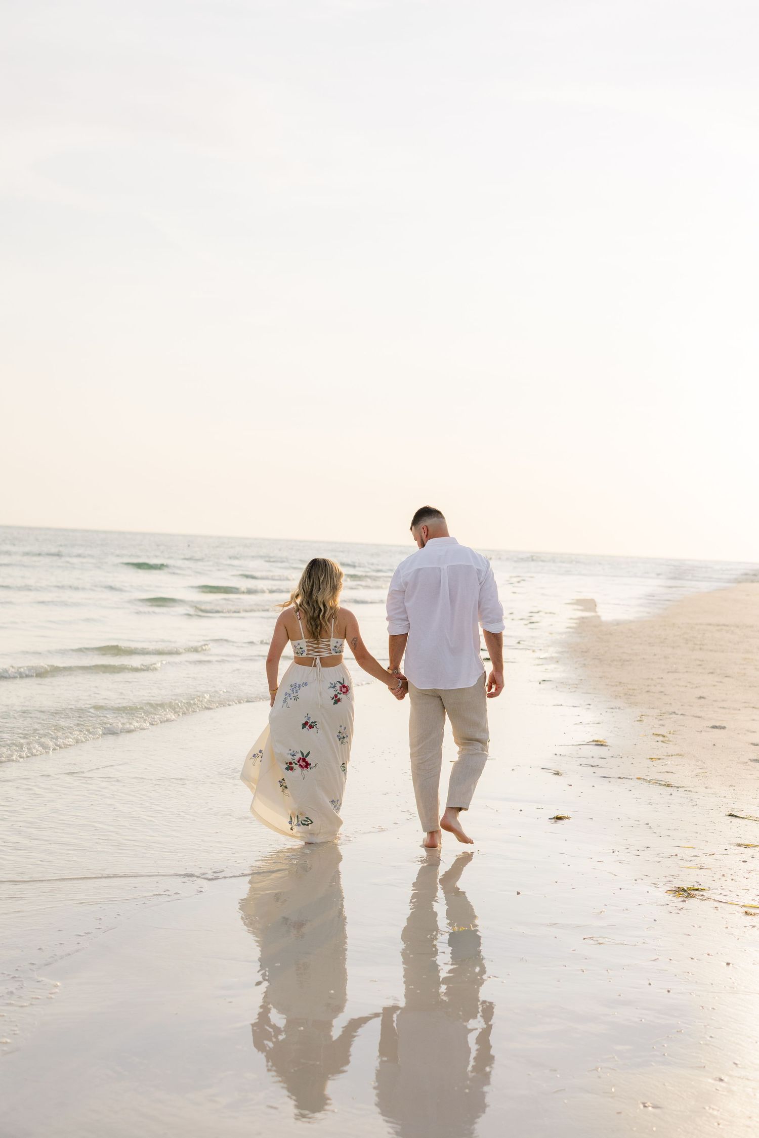Couple holding hands walking barefoot on a sandy beach at sunset, woman in floral dress, man in white shirt.