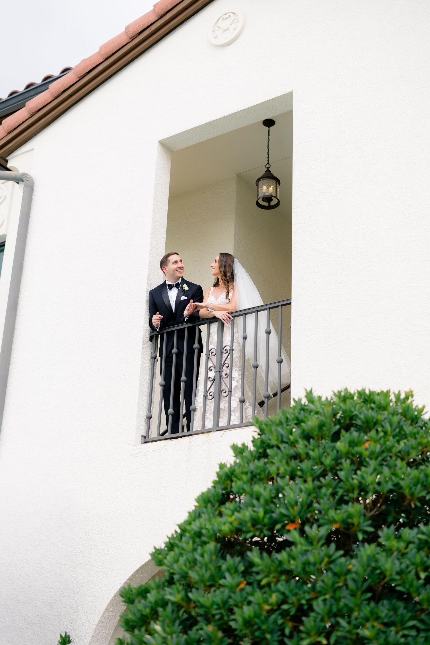 Bride and groom share a romantic moment on an elegant white balcony with wrought iron railing at their wedding venue.