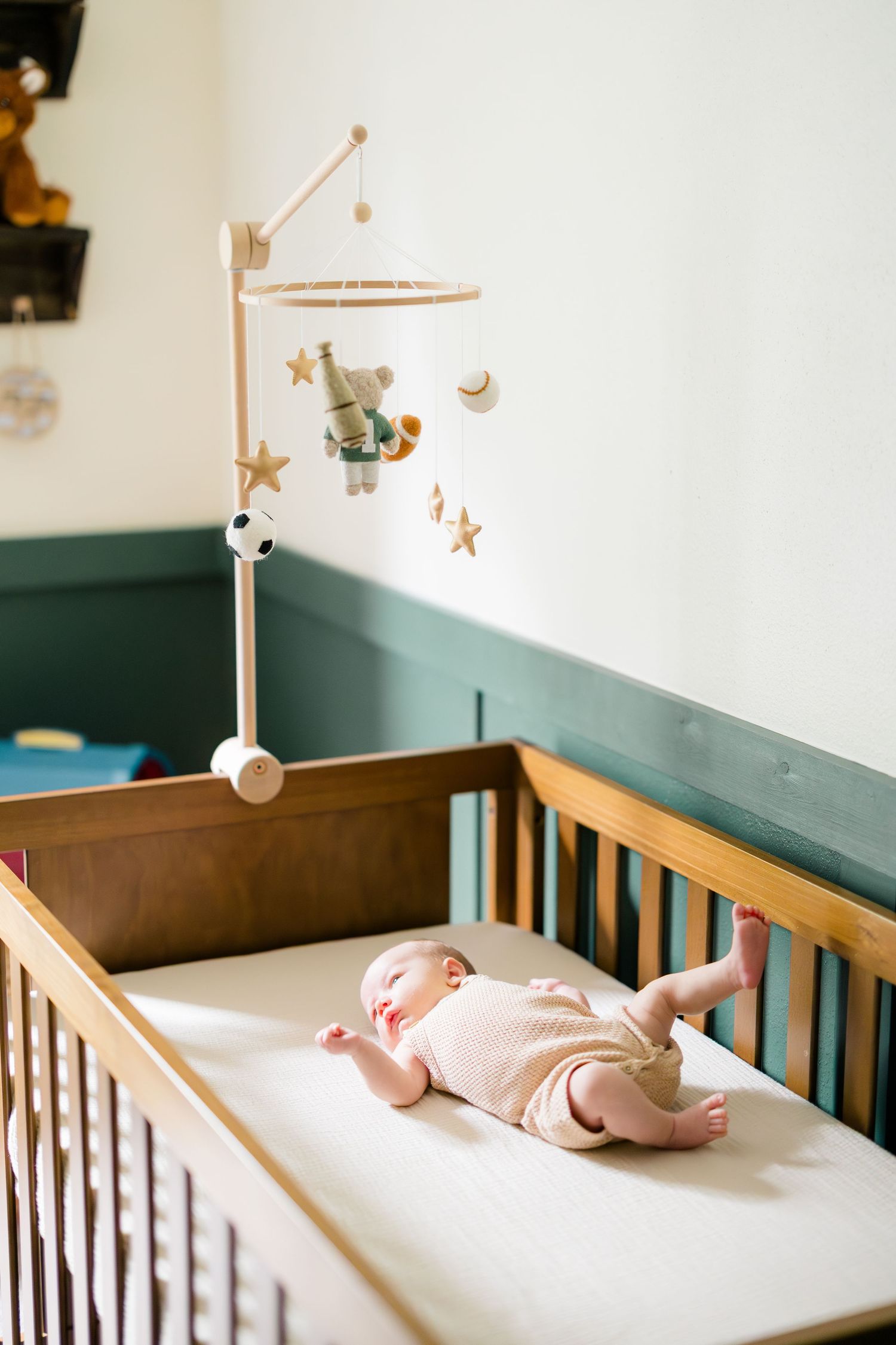 Newborn baby lying in wooden crib with star mobile toy in nursery room with green accent wall.