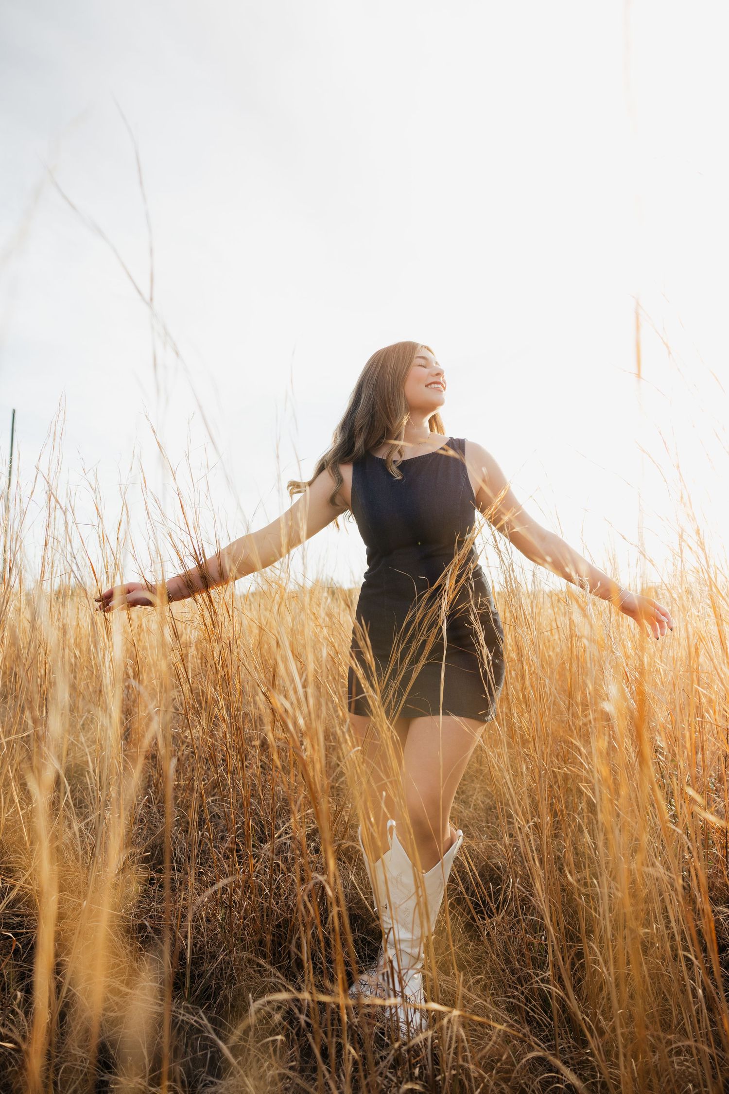 Young woman in black dress and white boots joyfully walks through golden wheat field with arms outstretched.