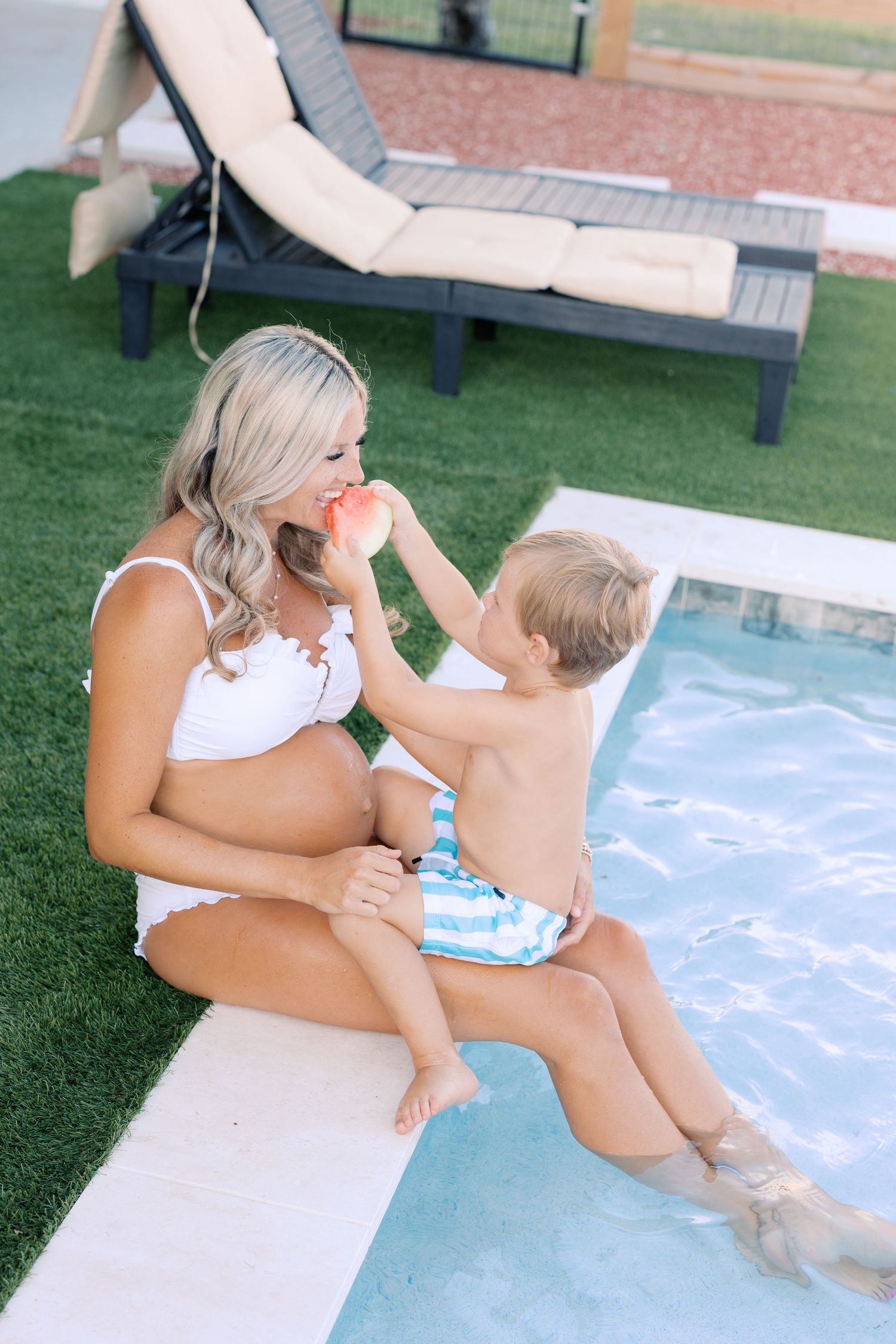Pregnant woman in white bikini sitting poolside while toddler boy feeds her watermelon on sunny day.