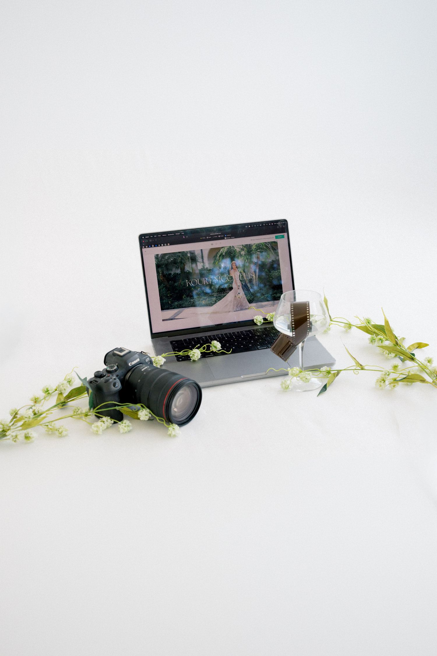 Camera and laptop with wedding photo editing software, surrounded by delicate white flowers on white background.