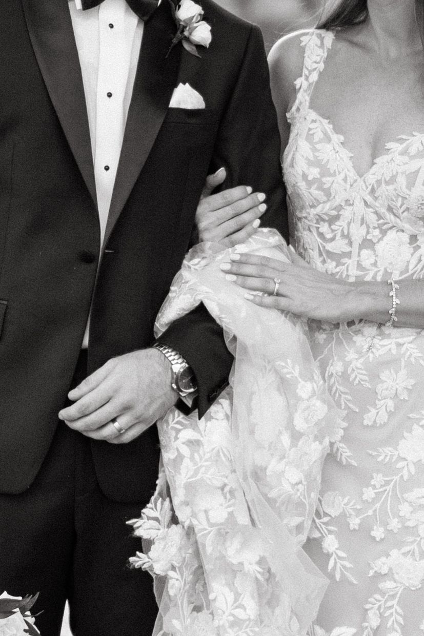 Black and white photo of bride and groom embracing, showing wedding rings, floral lace gown, and tuxedo details.