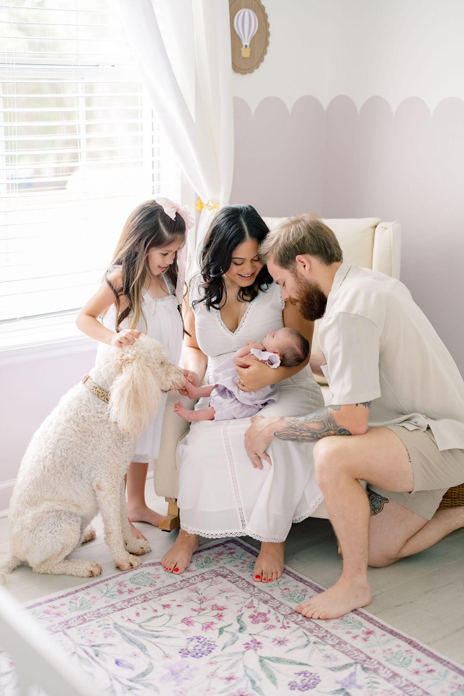 Family with newborn baby, toddler girl, and fluffy dog gathered together in a soft pastel nursery room.