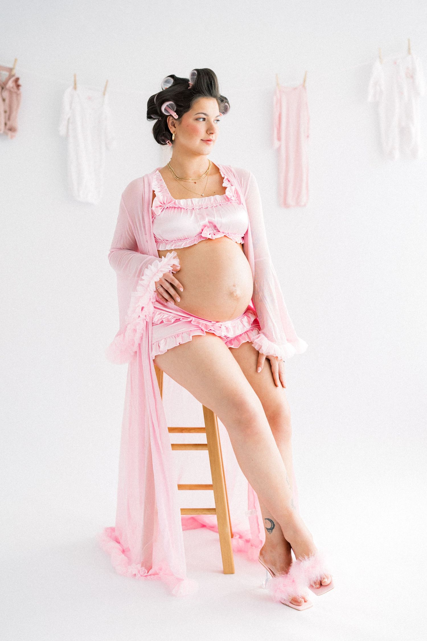Pregnant woman in pink lingerie and robe with hair rollers sits on wooden stool against white backdrop with hanging baby clothes.