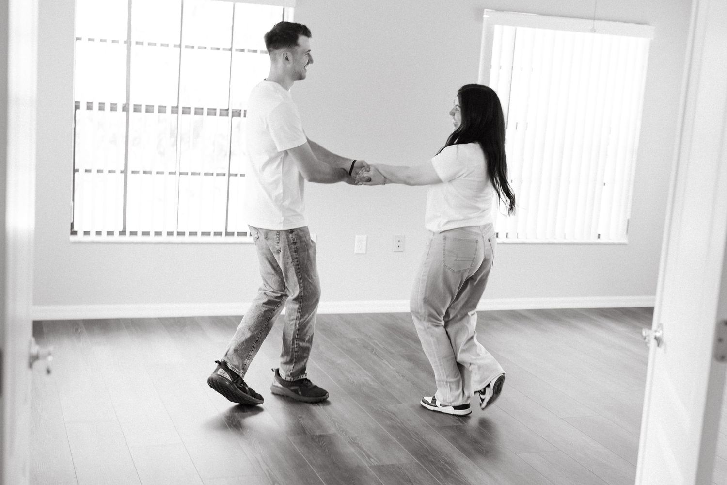 A couple dances together joyfully in an empty room with hardwood floors and large windows in black and white.