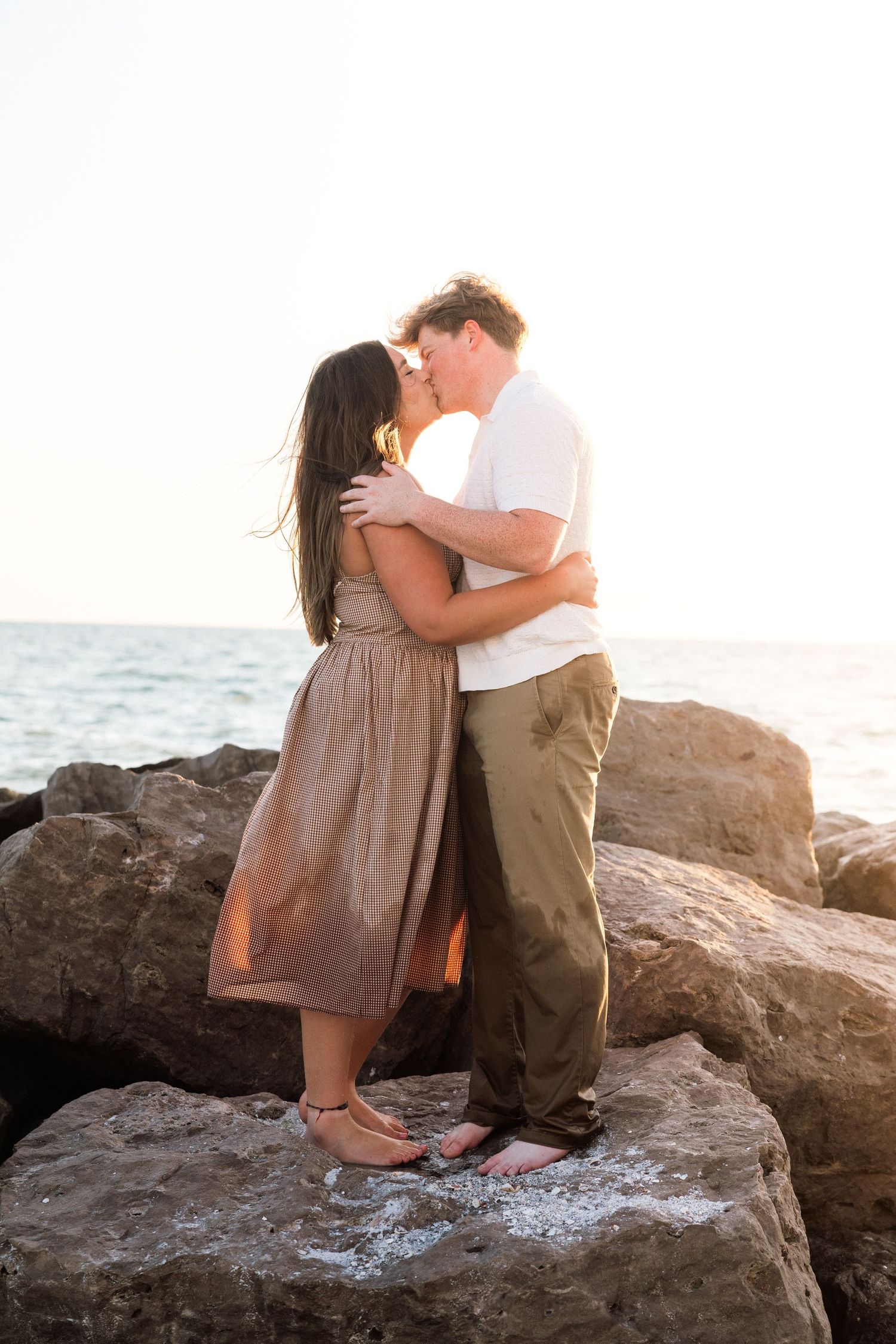 Couple kissing on rocky shoreline at sunset, woman in tan dress, man in white shirt and green pants.