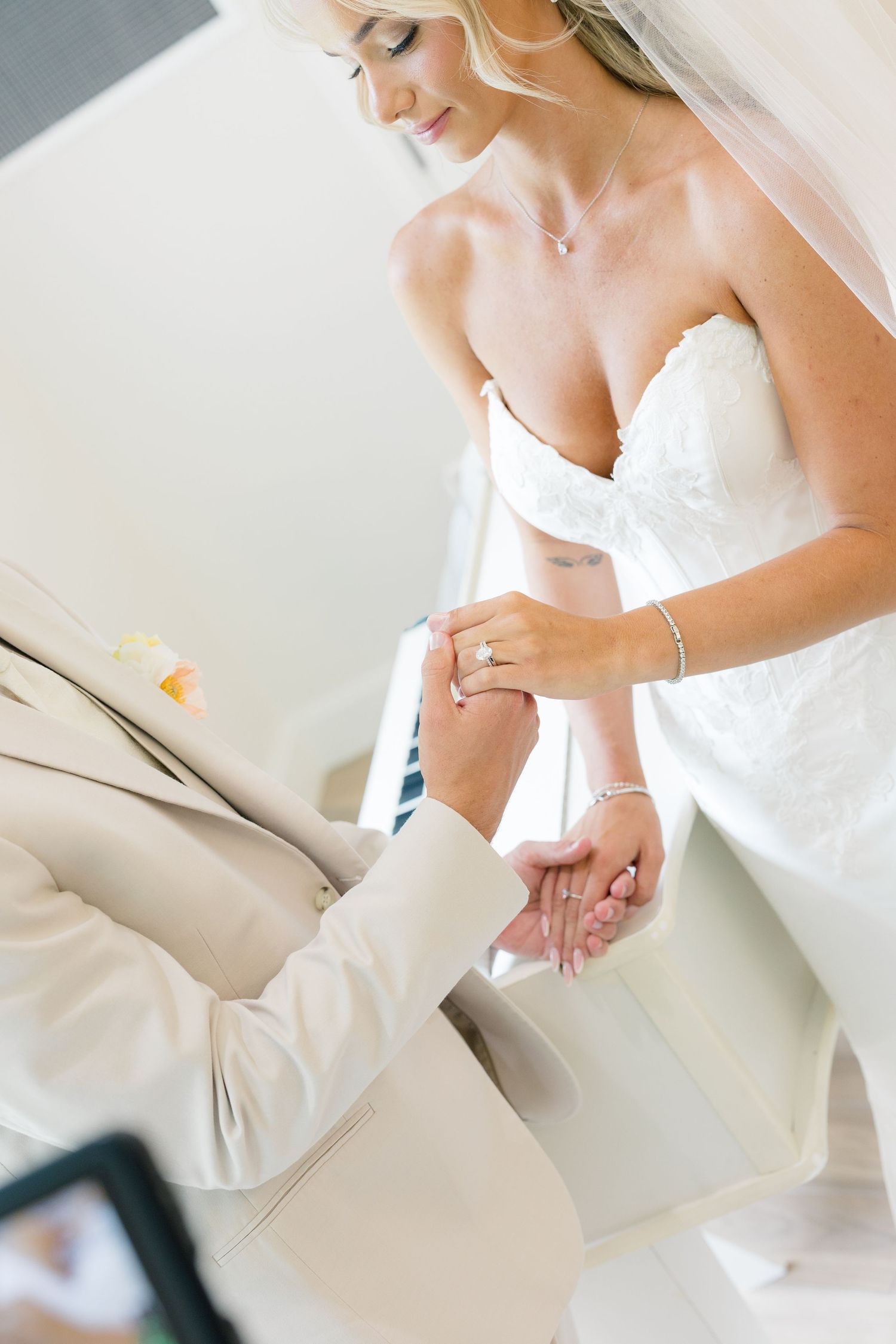 Bride in strapless lace gown and veil exchanges rings with groom in beige suit during wedding ceremony.