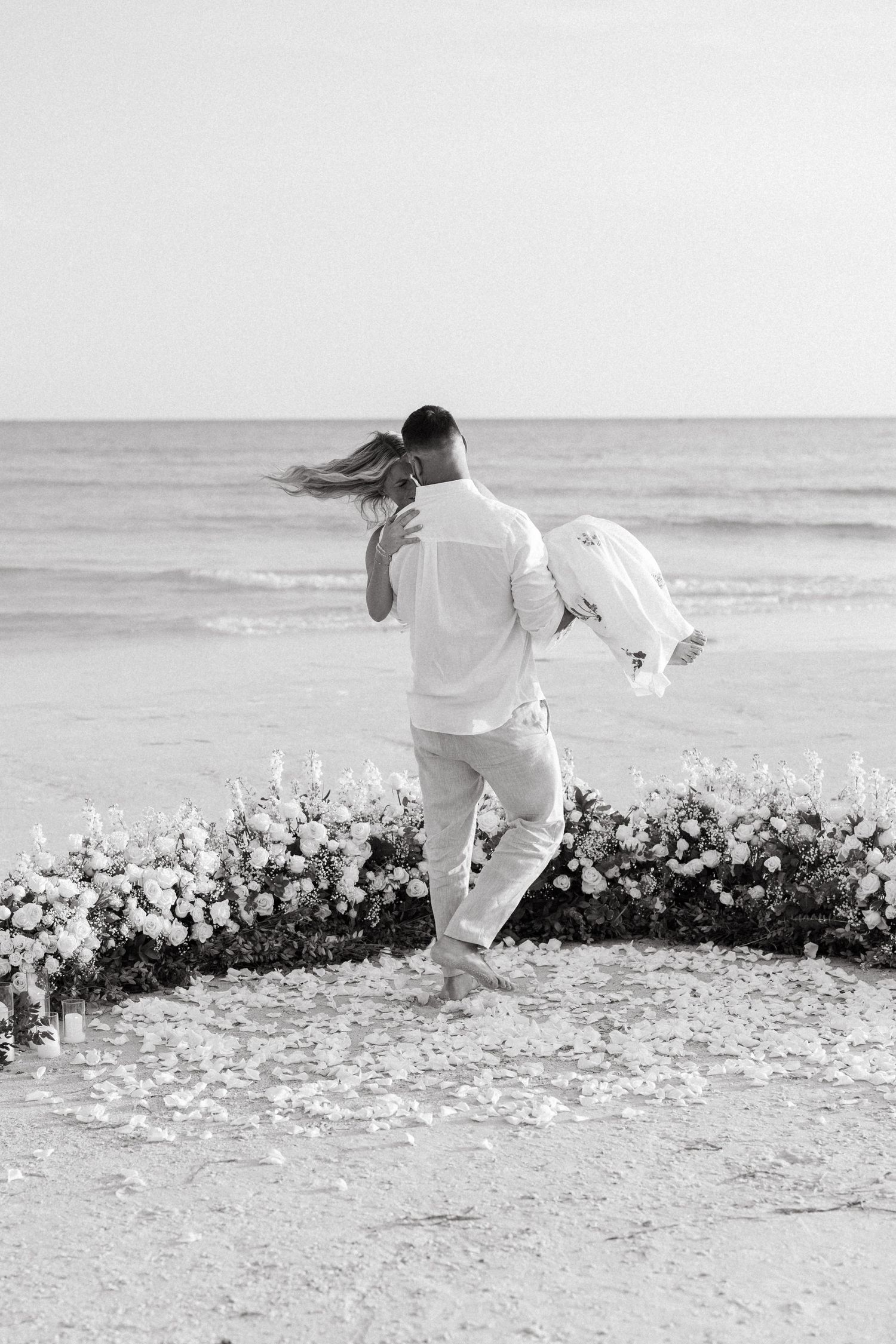 Black and white beach proposal photo of couple embracing in floral circle arrangement with ocean backdrop.