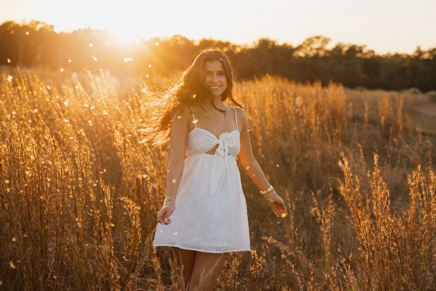 Young woman in white dress walking through golden field at sunset with warm sunlight glowing behind her.