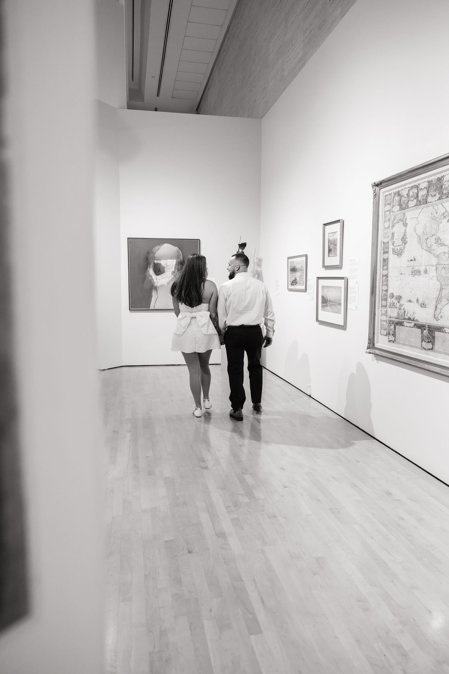 Couple walking hand in hand through a modern art gallery, viewing paintings on white walls, black and white photo.