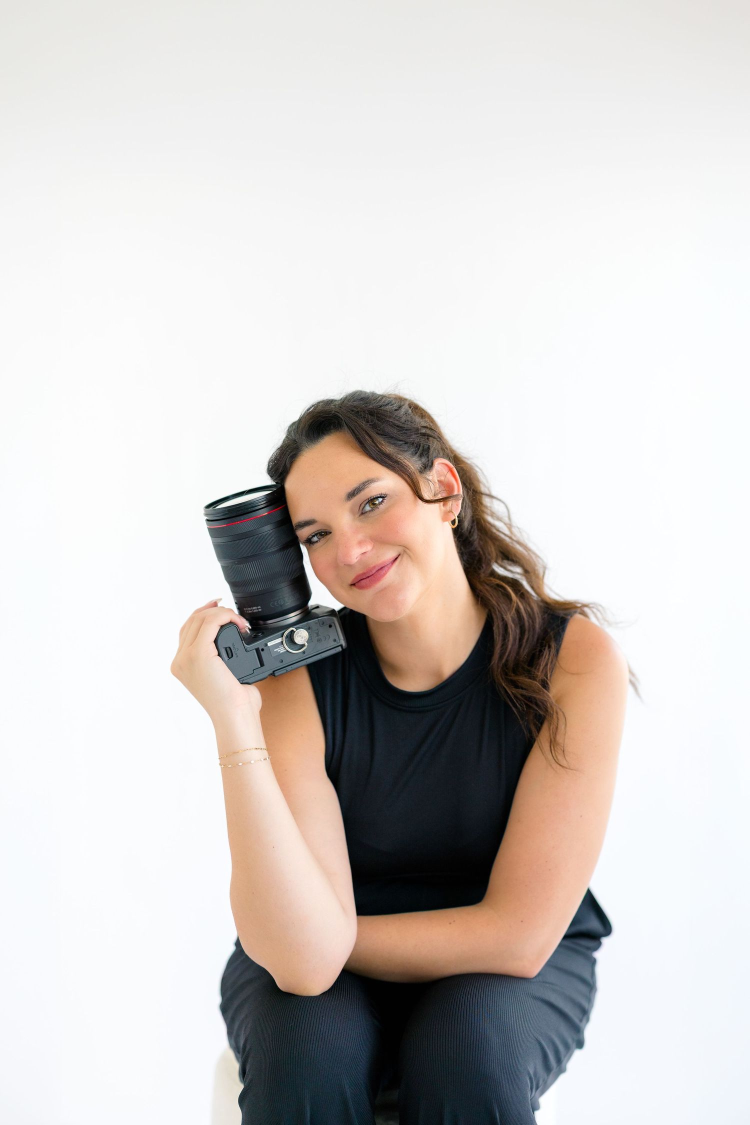 A smiling woman in a black outfit holds a DSLR camera beside her face against a white background.