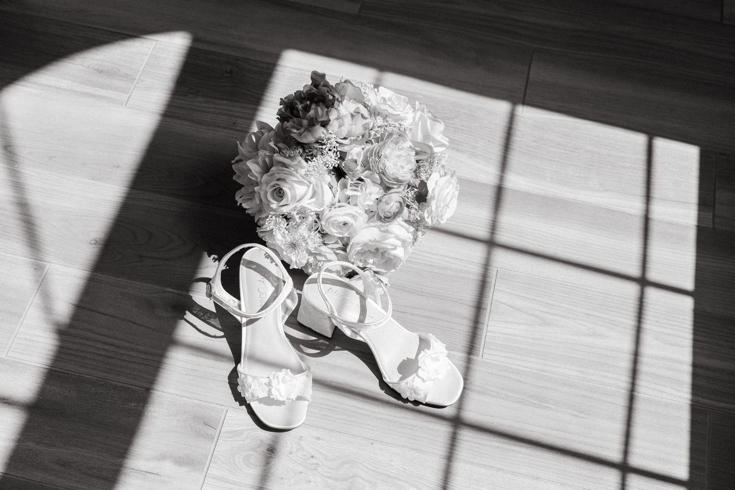 Black and white photo of bridal heels and a rose bouquet on a wooden floor with dramatic light shadows.