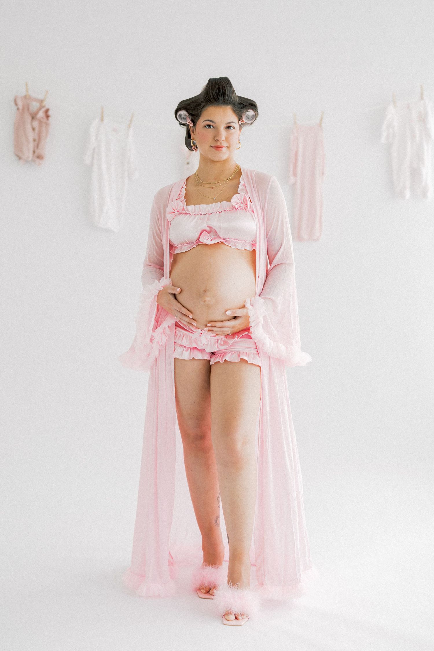 Pregnant woman in pink lingerie set and sheer robe with hair curlers, posing in white studio with baby clothes.