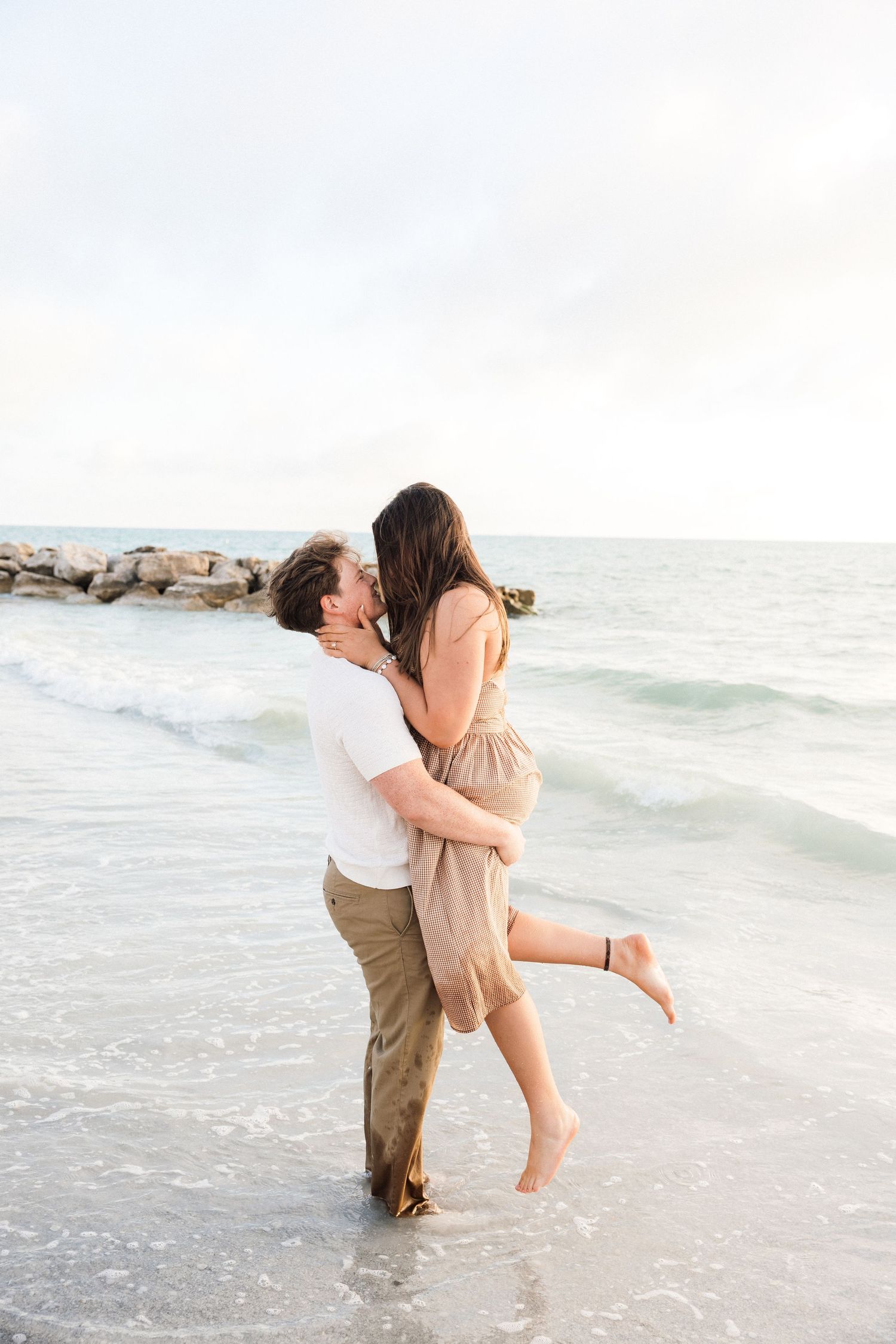 Couple embracing and kissing on a beach shore, man lifting woman in a romantic moment with ocean waves in the background.