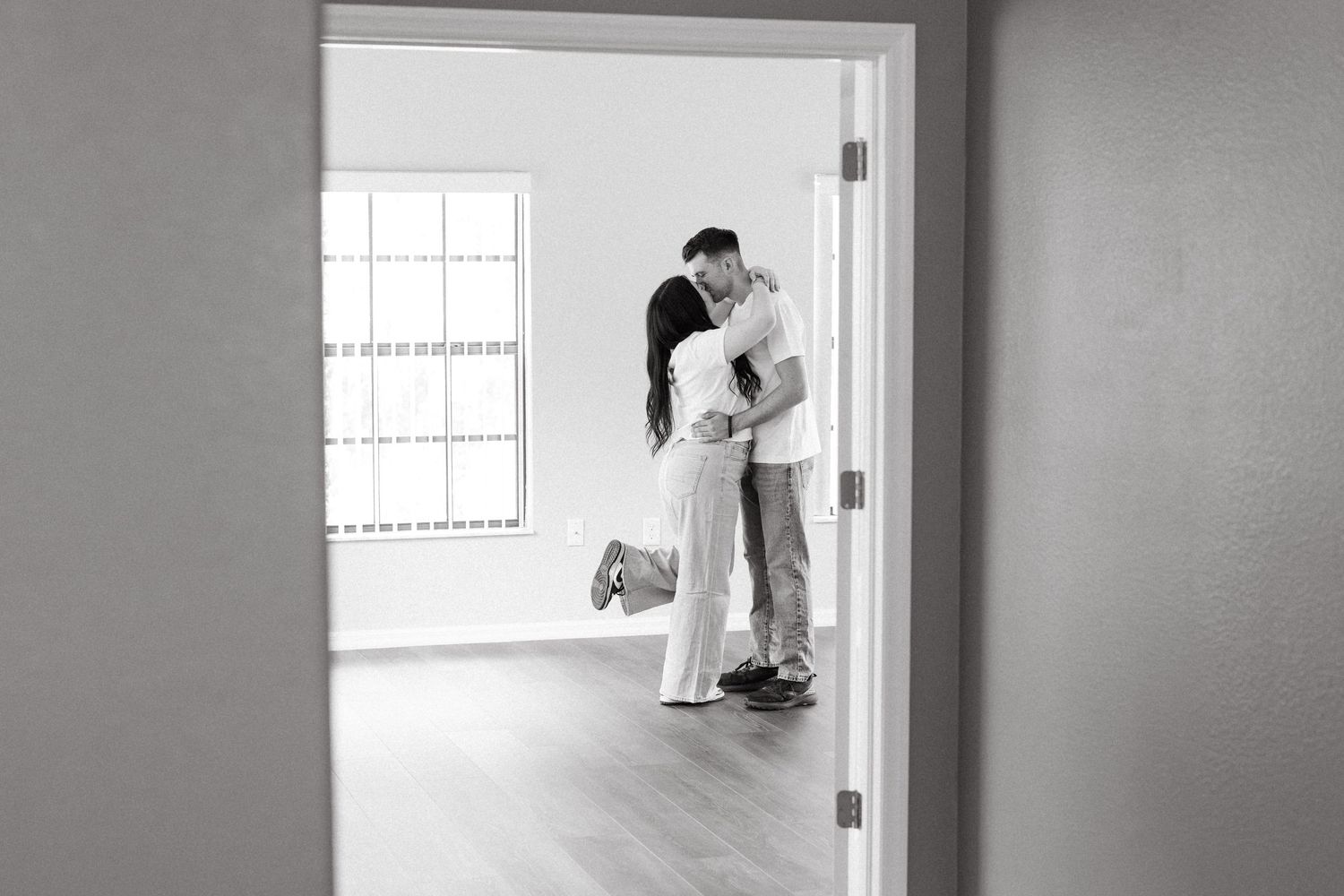 Black and white photo of a couple sharing a romantic kiss in an empty room, viewed through a doorway.
