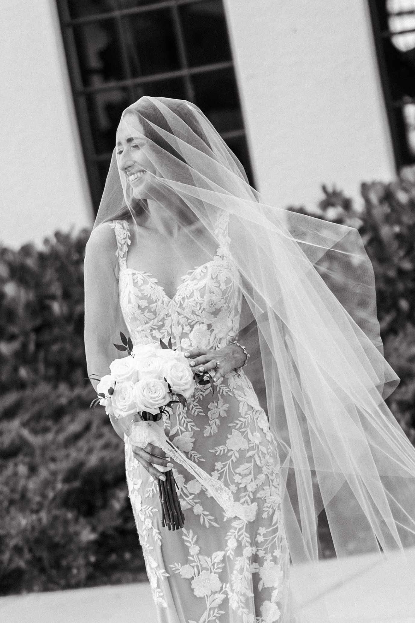 Black and white photo of a bride in lace gown holding white bouquet, veil flowing in the wind outdoors.