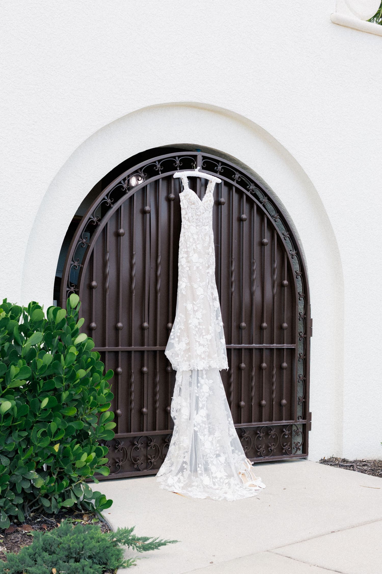 Elegant lace wedding dress hanging on ornate iron arched door of white stucco building with lush greenery.