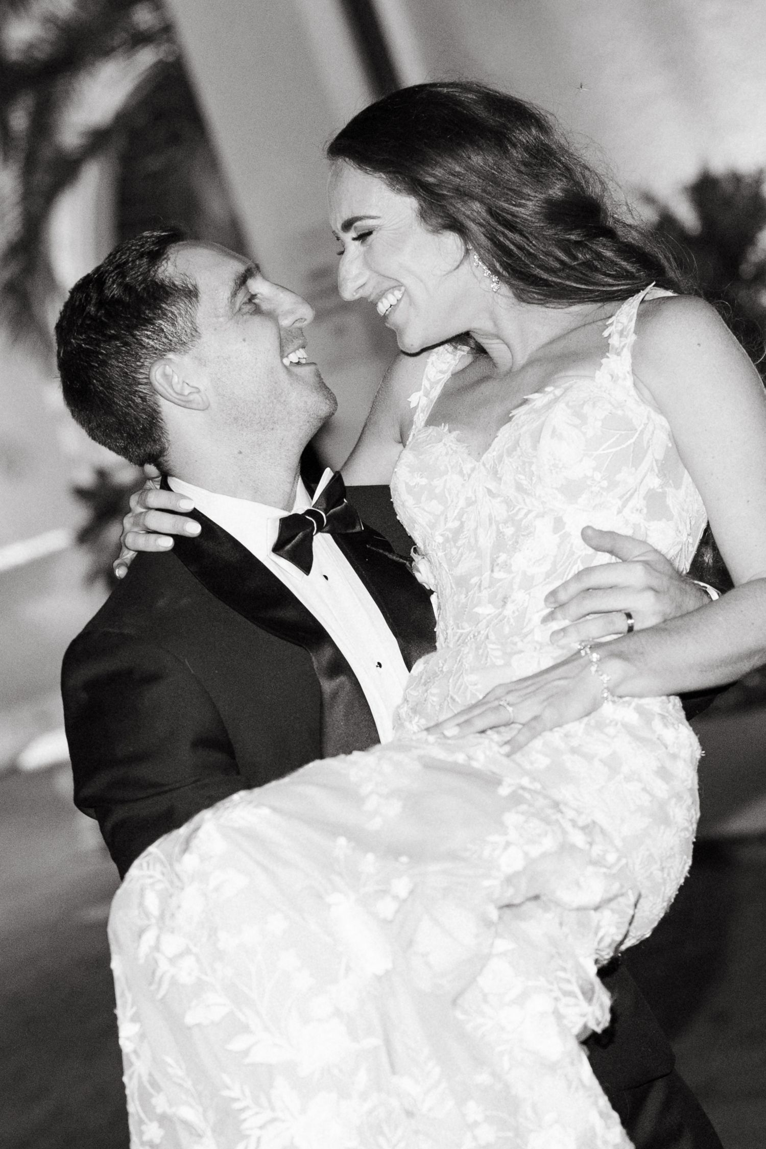 Black and white photo of a groom lifting his bride in a wedding gown, both laughing joyfully.