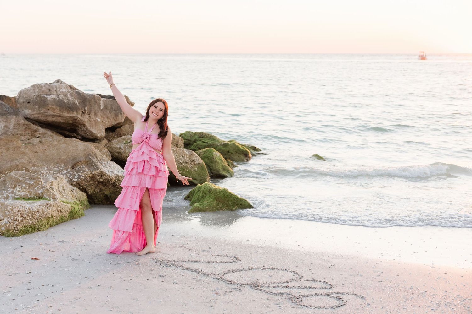 Woman in pink ruffled gown poses on beach near rocks with 2023 written in sand at sunset.