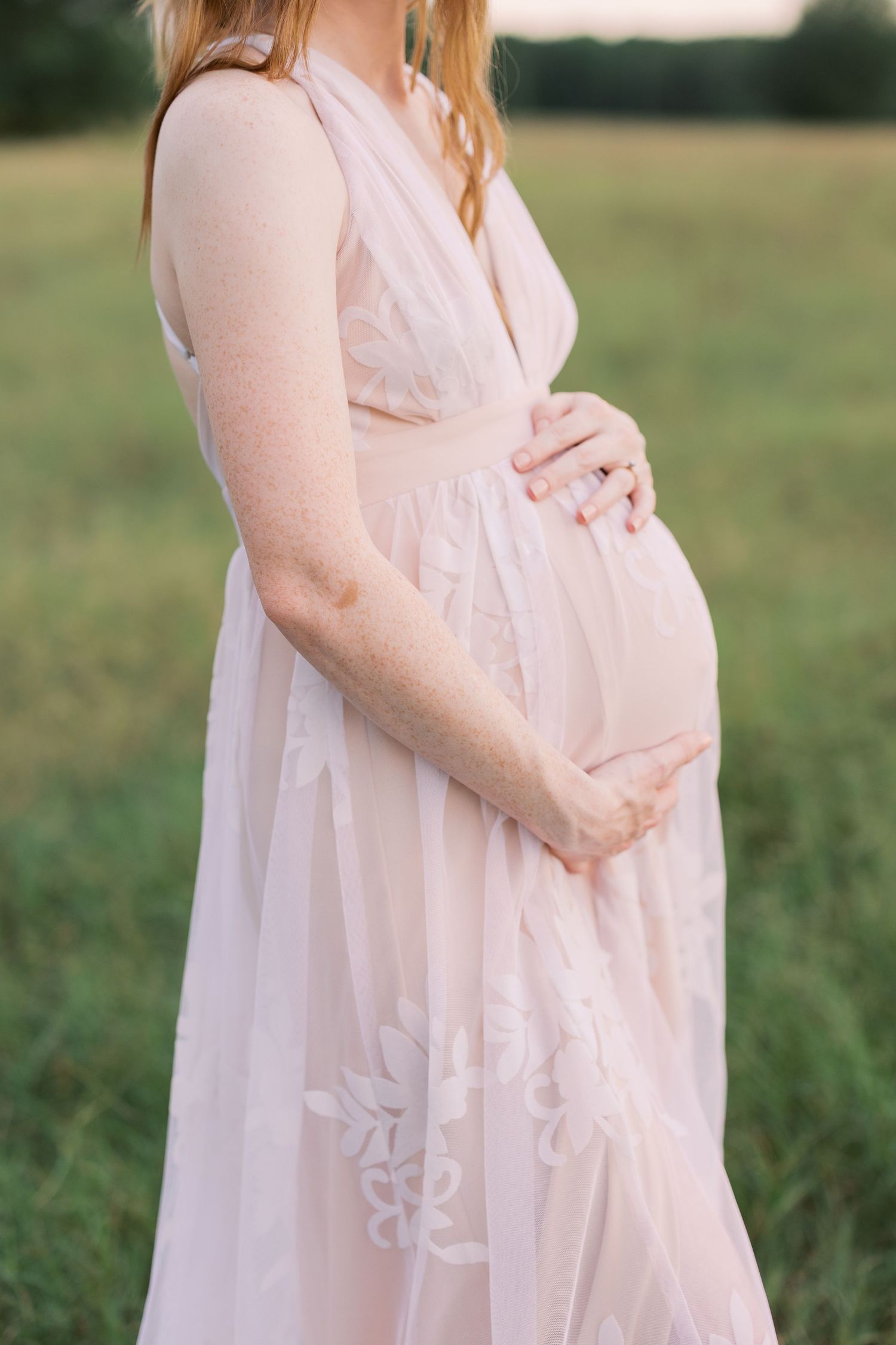 Florida maternity portrait of pregnant mother in blush pink lace gown cradling her baby bump in a green field.