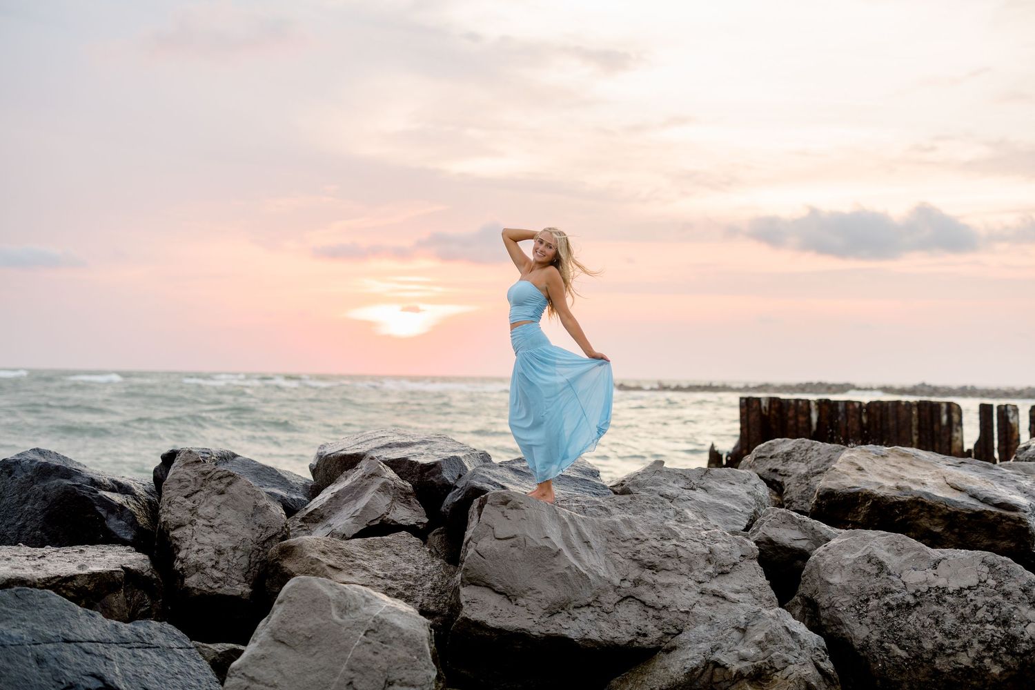 Senior in flowing blue dress on the rocky shoreline at St. Pete Beach during sunset.