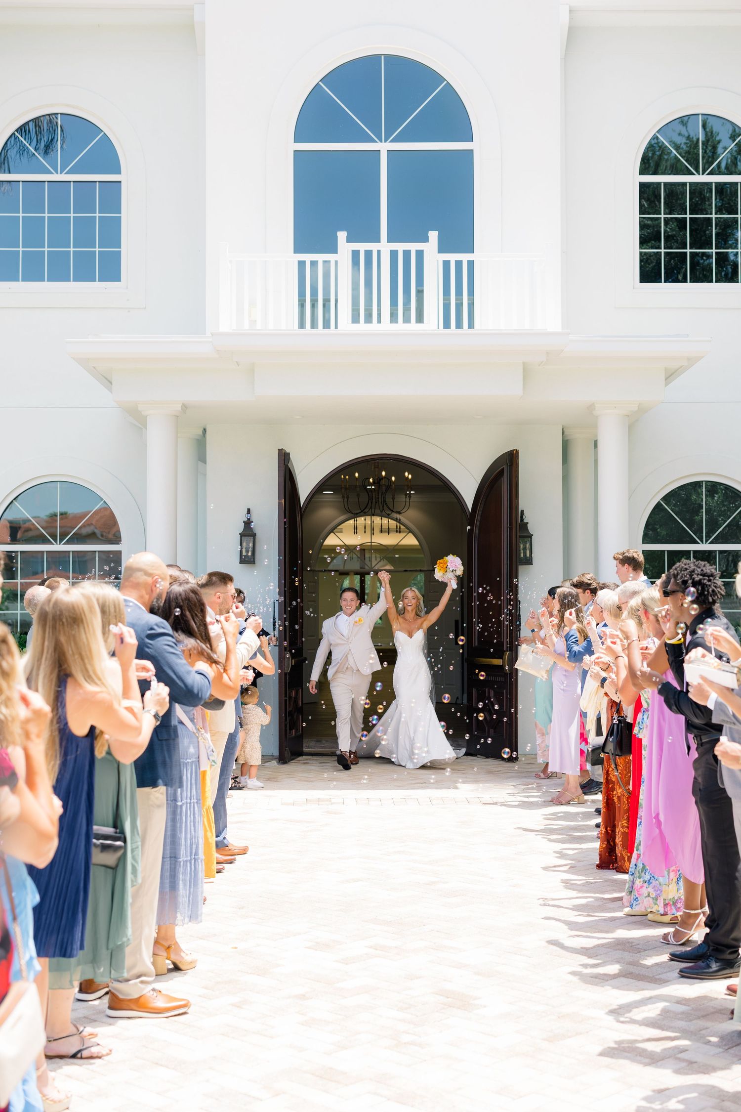 Bride and groom exit white wedding venue through arched doors as guests line up to celebrate with confetti.