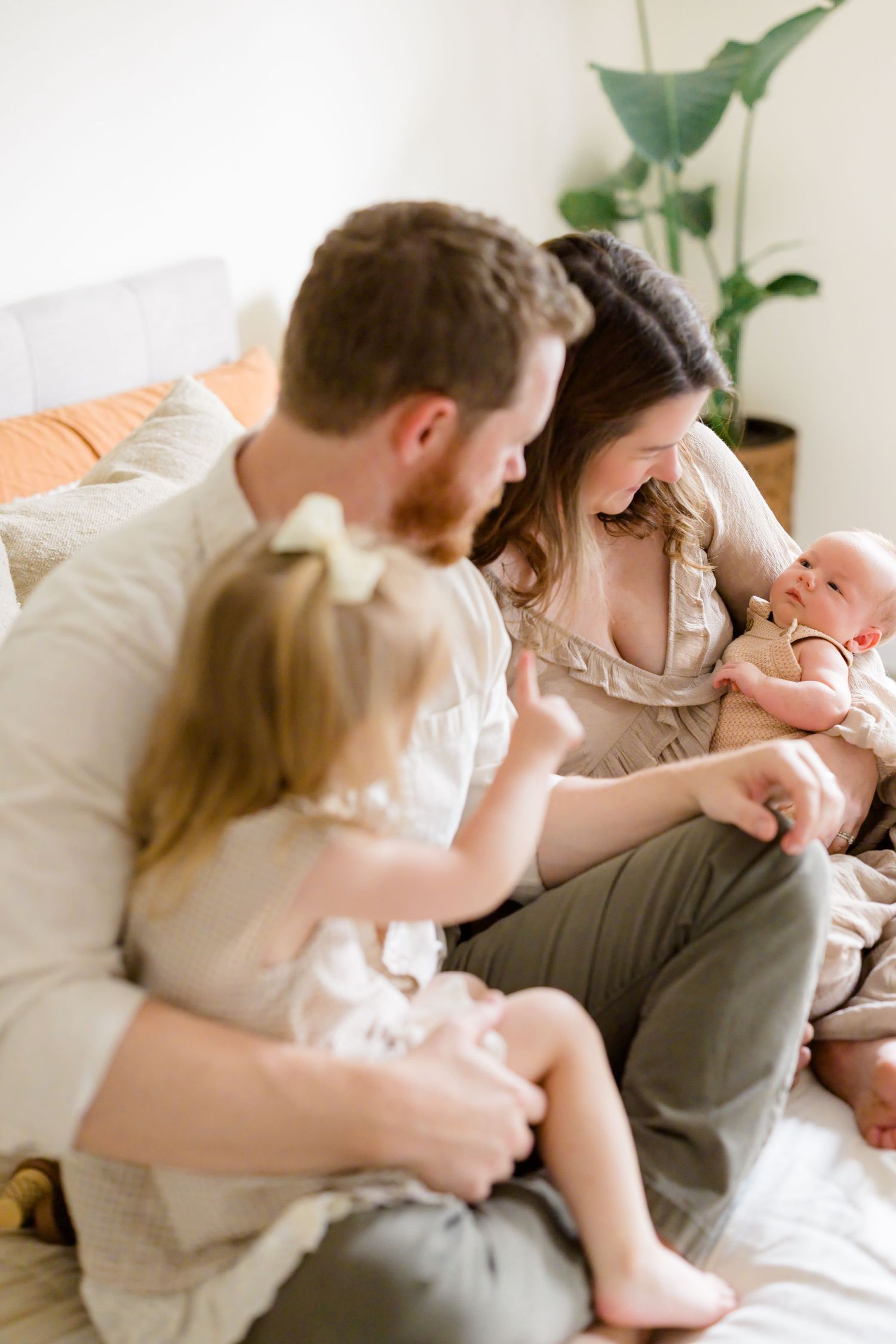 Family of four sitting on bed, parents admiring newborn baby while toddler daughter reaches out to touch sibling.