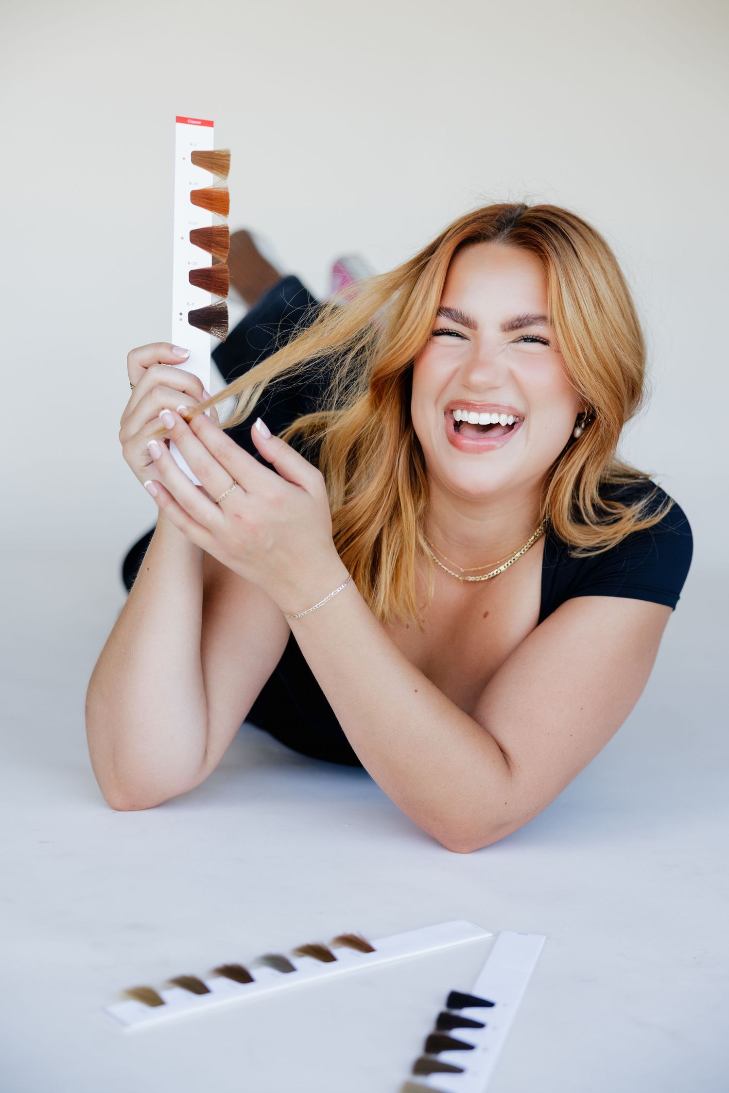 Smiling woman lying down holding hair color swatches, with additional color samples displayed on white surface nearby.