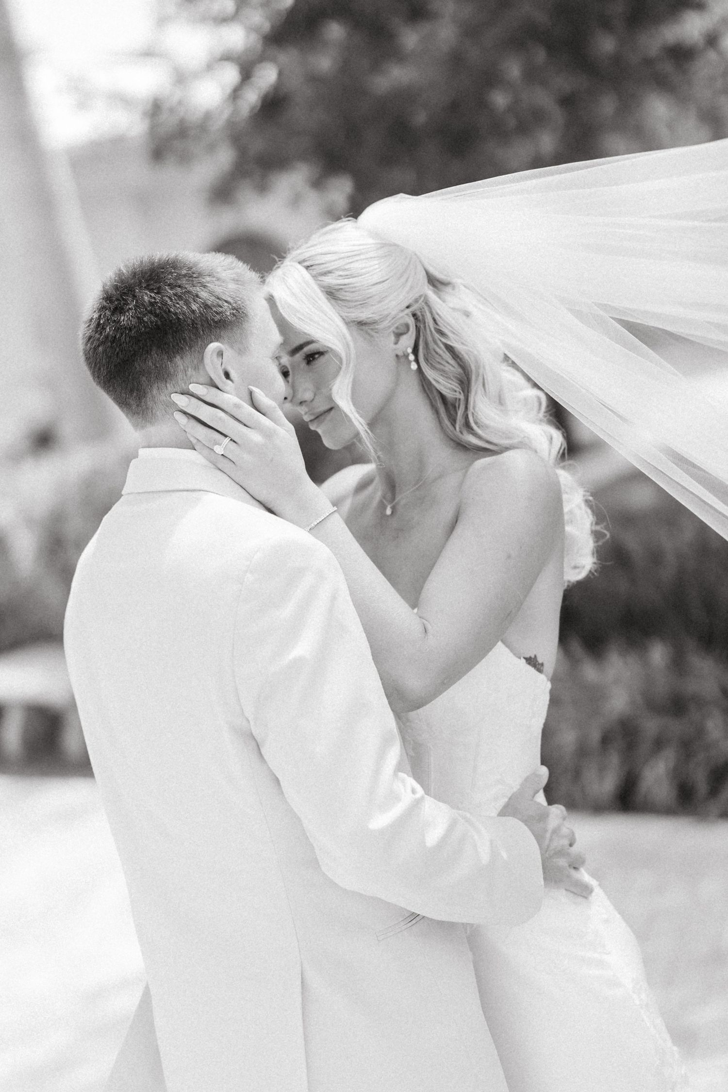 Black and white photo of bride and groom sharing romantic moment, veil flowing in breeze.