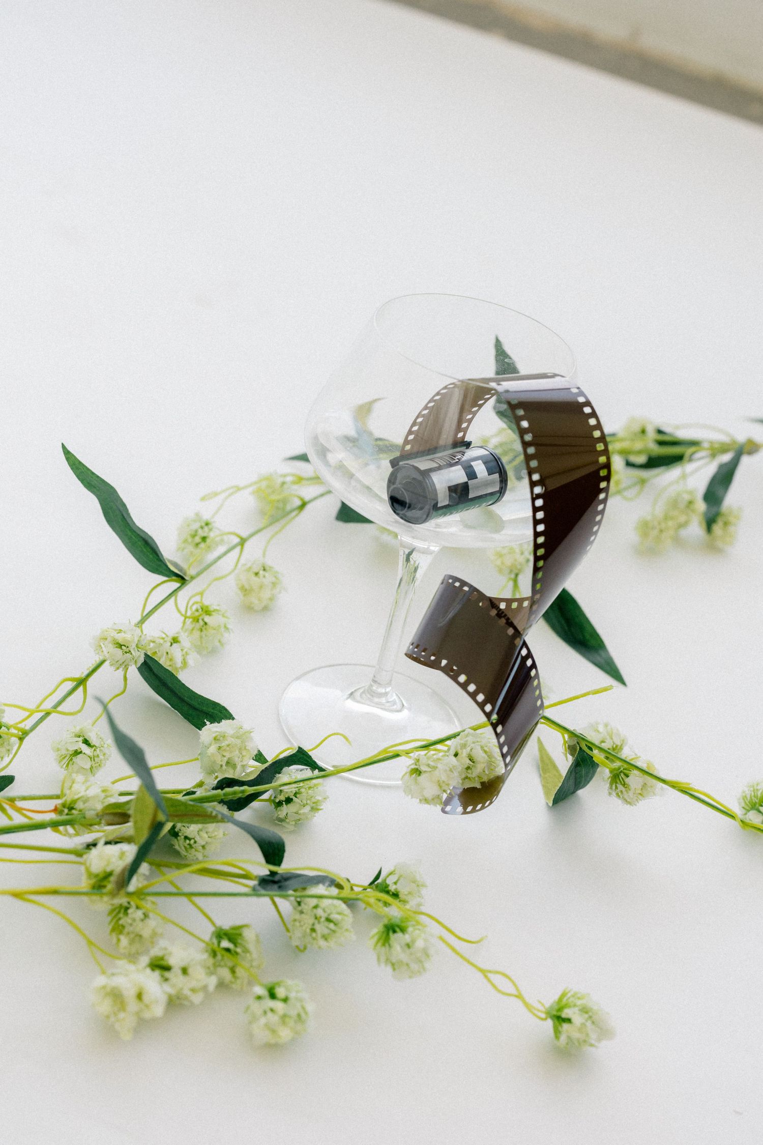 A clear wine glass with vintage film strip surrounded by delicate white wildflowers on a white surface.
